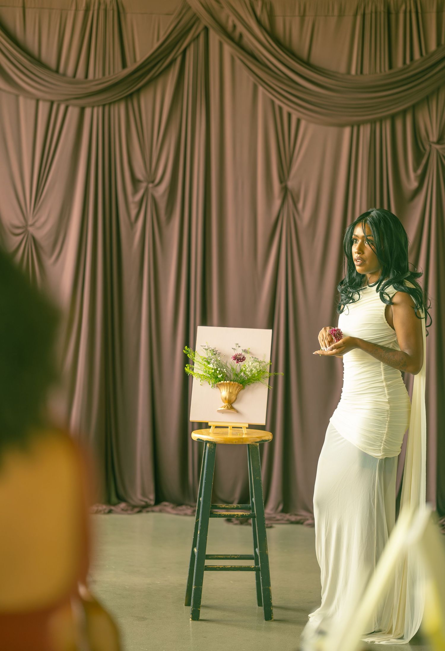 Woman in a white gown speaks beside a framed floral painting on a wooden stool, with draped mauve curtains behind her.