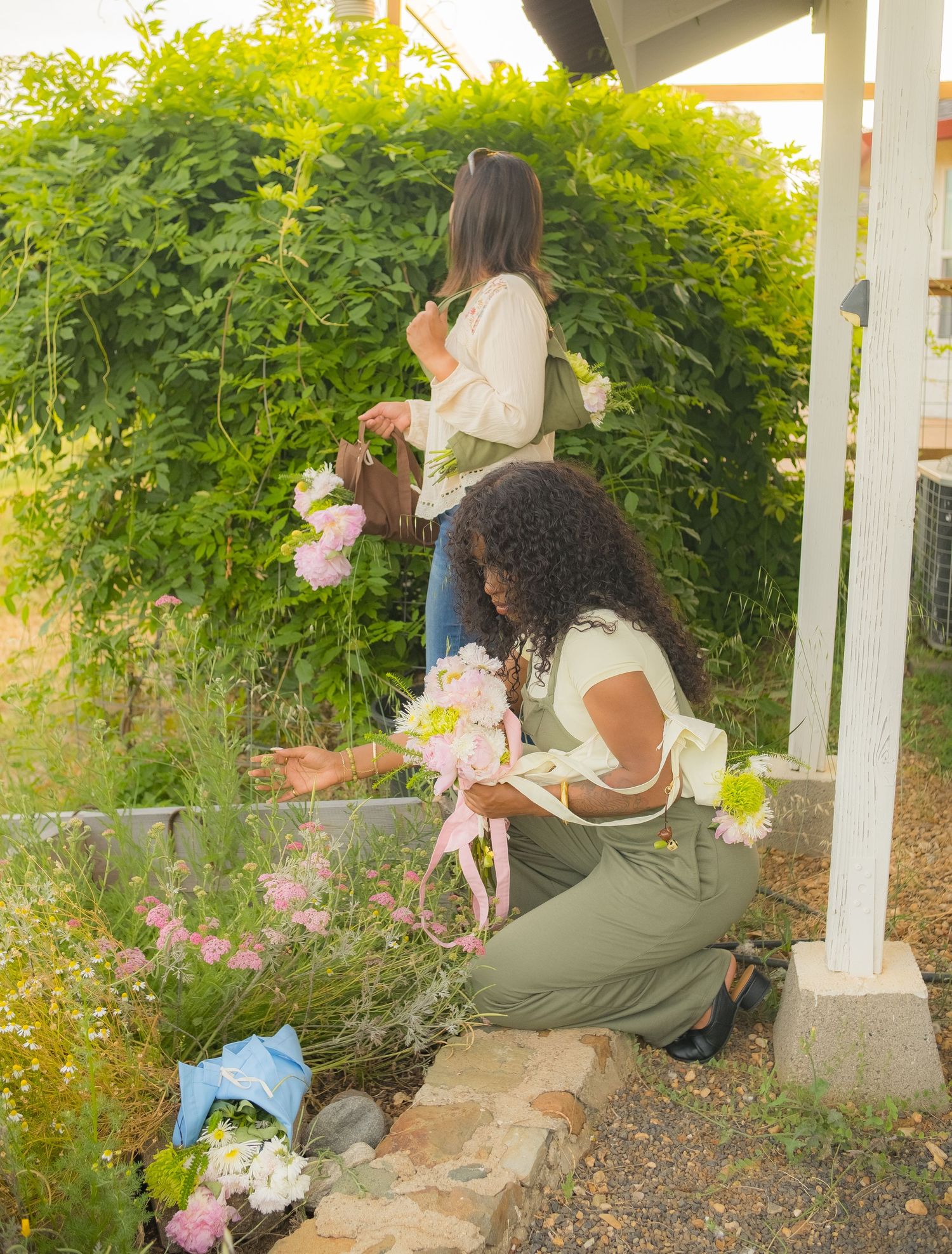 Two women arranging fresh pink flowers in a lush garden, one kneeling and one standing near greenery.