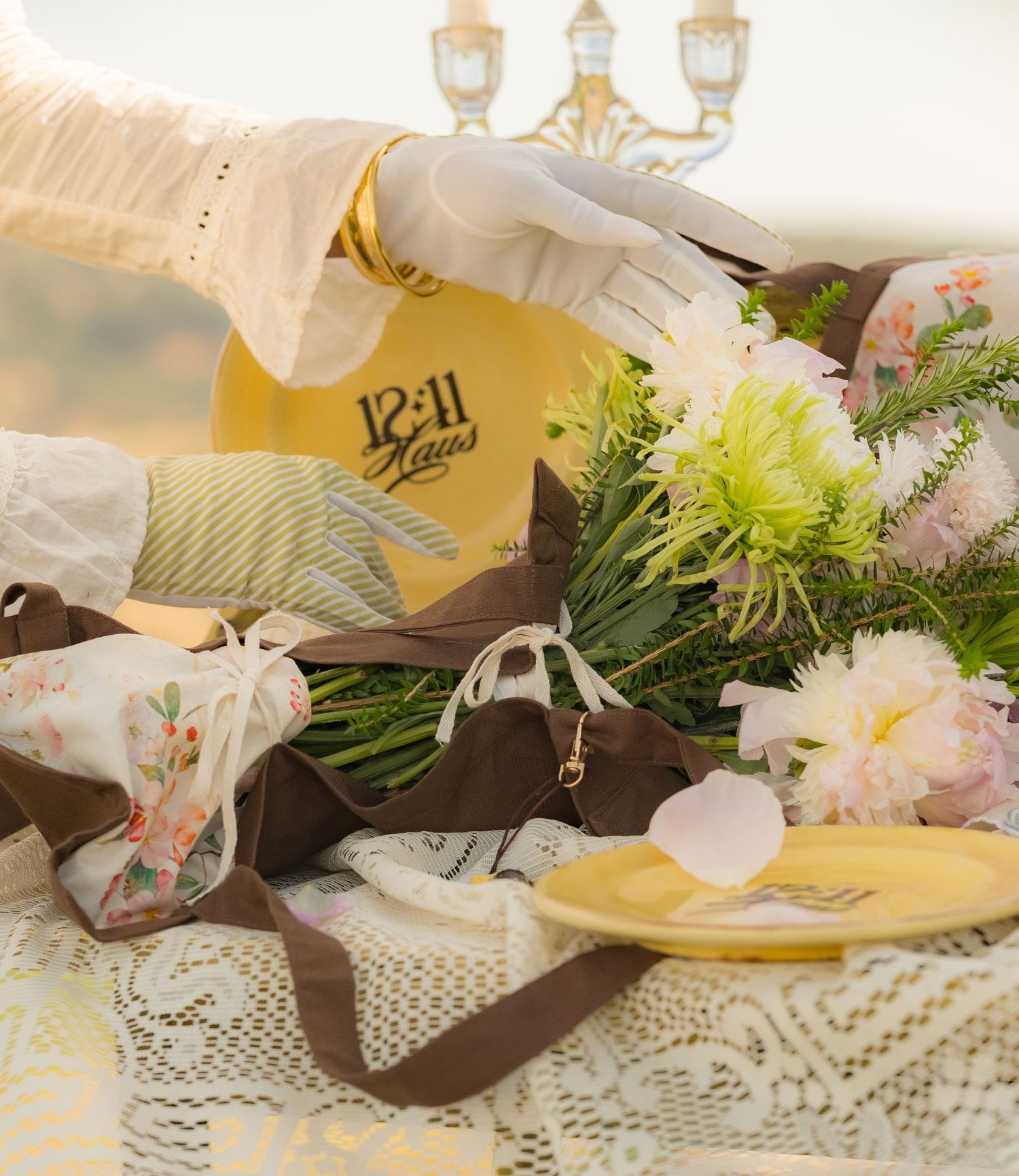 Elegant table setting with floral arrangement, gold clock, brown bag, and white gloved hand holding a yellow item.