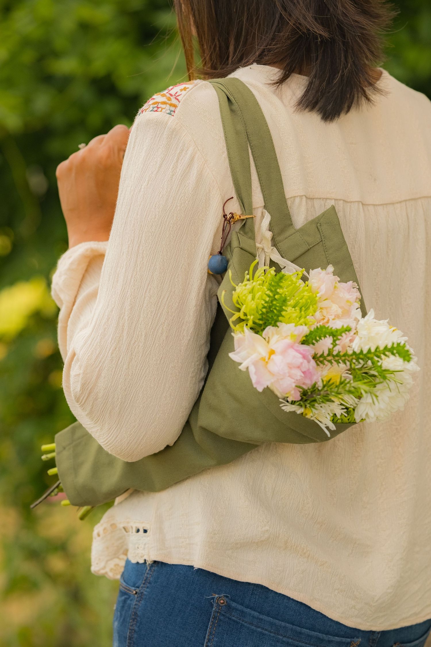 Woman carrying a green canvas tote bag filled with fresh pink and white flowers outdoors.