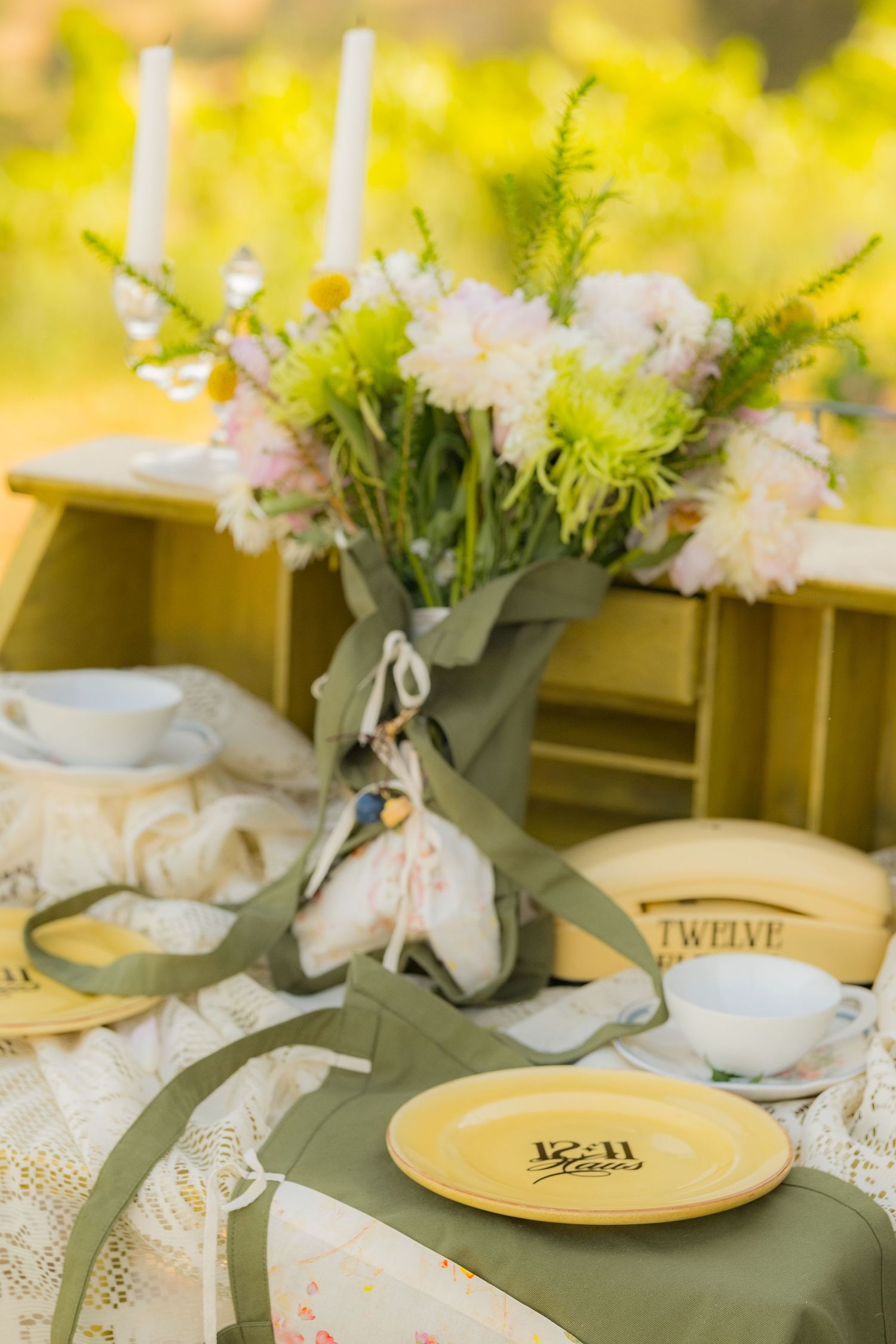Elegant outdoor table setting with yellow plates, lace tablecloth, candles, and pink and green floral centerpiece.
