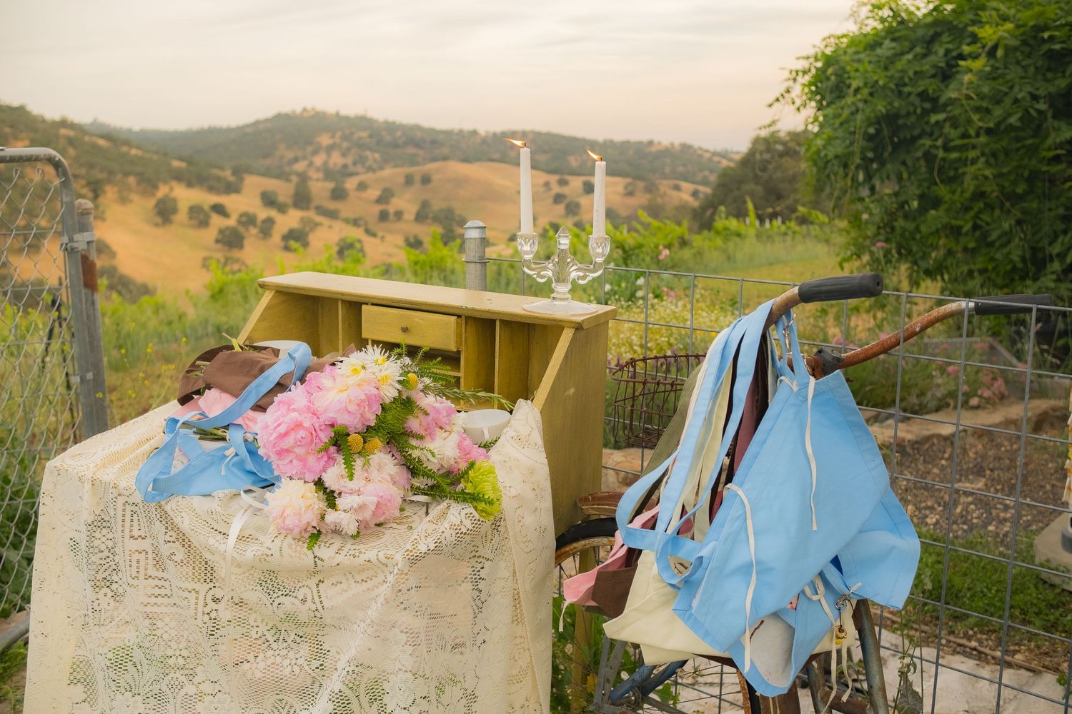 Vintage yellow dresser with pink flowers and blue bag on fence, overlooking scenic rolling hills at sunset.