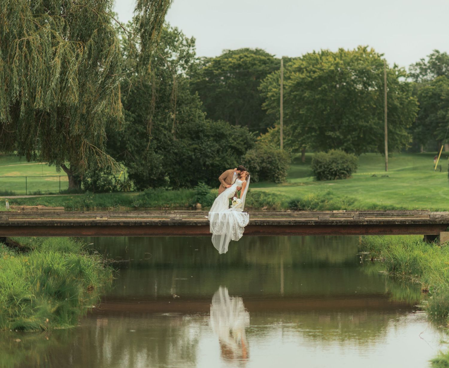 Bride in a flowing white gown sits on a wooden bridge over a calm pond, reflected in the water, with willow trees and a green park behind.