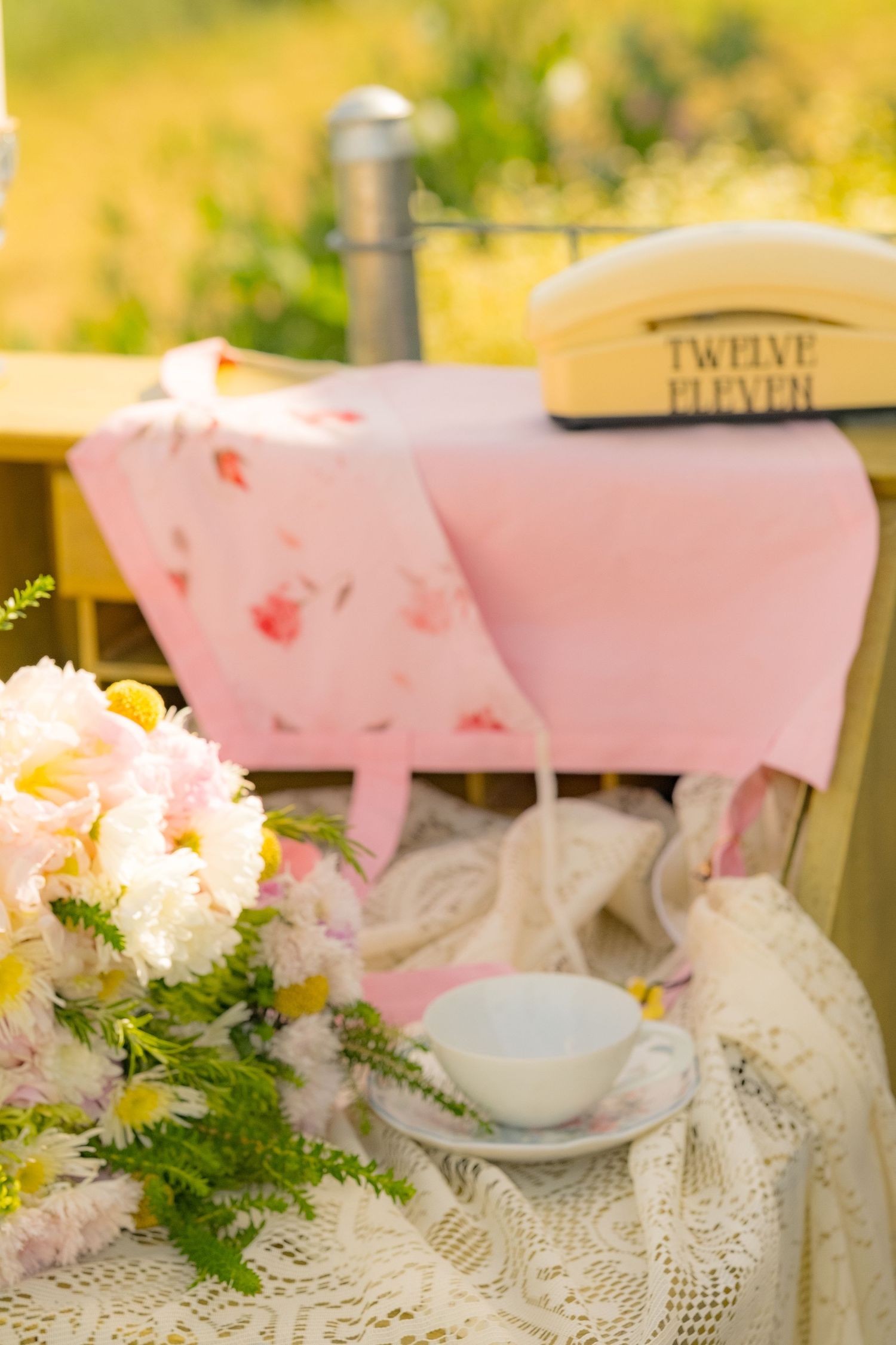 Vintage outdoor tea setting with white teacup, lace cloth, pink floral fabric, yellow flowers, and a retro phone on a sunny day.