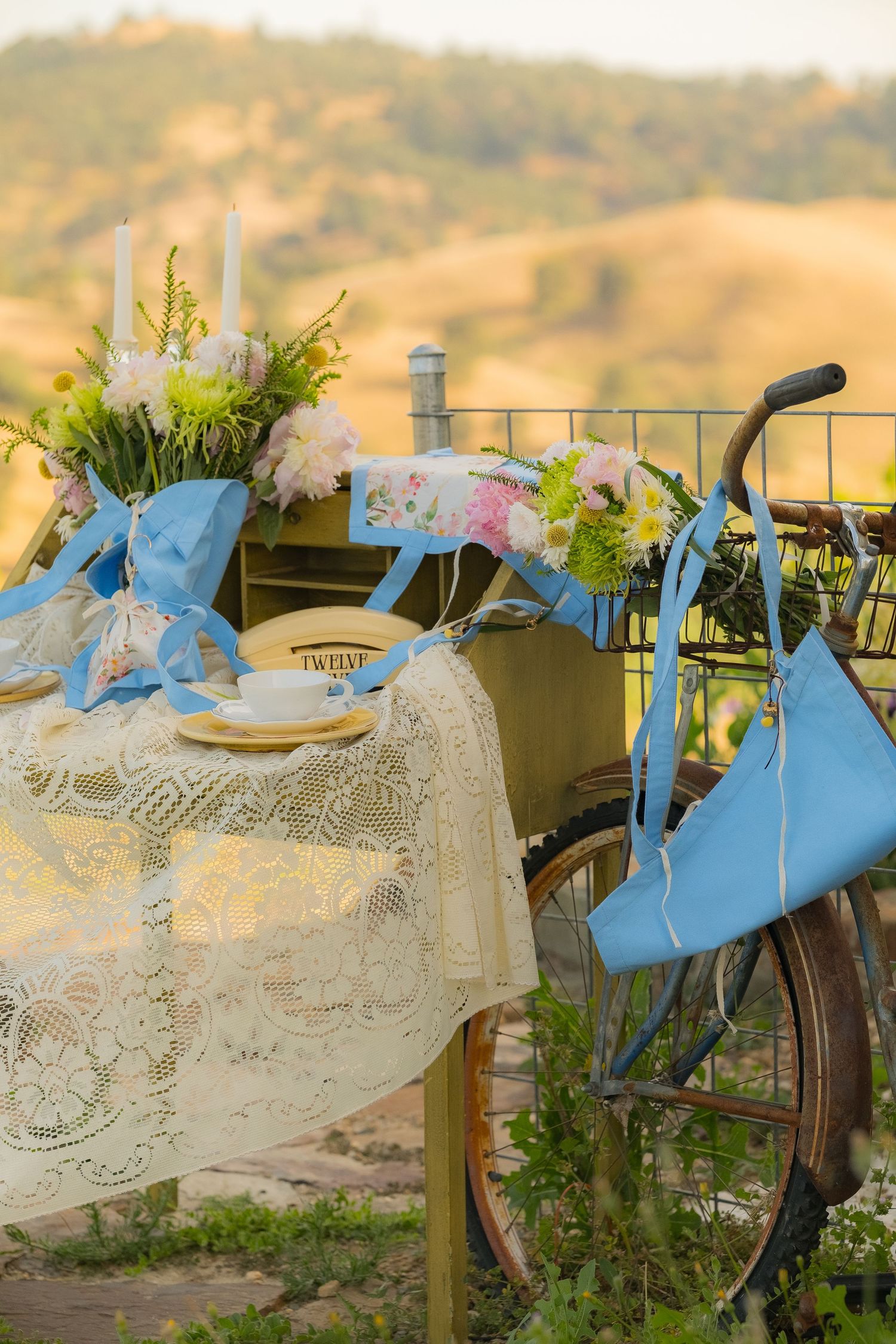Vintage bicycle decorated with blue ribbons beside a lace-covered table set with flowers and candles in a rustic outdoor setting.