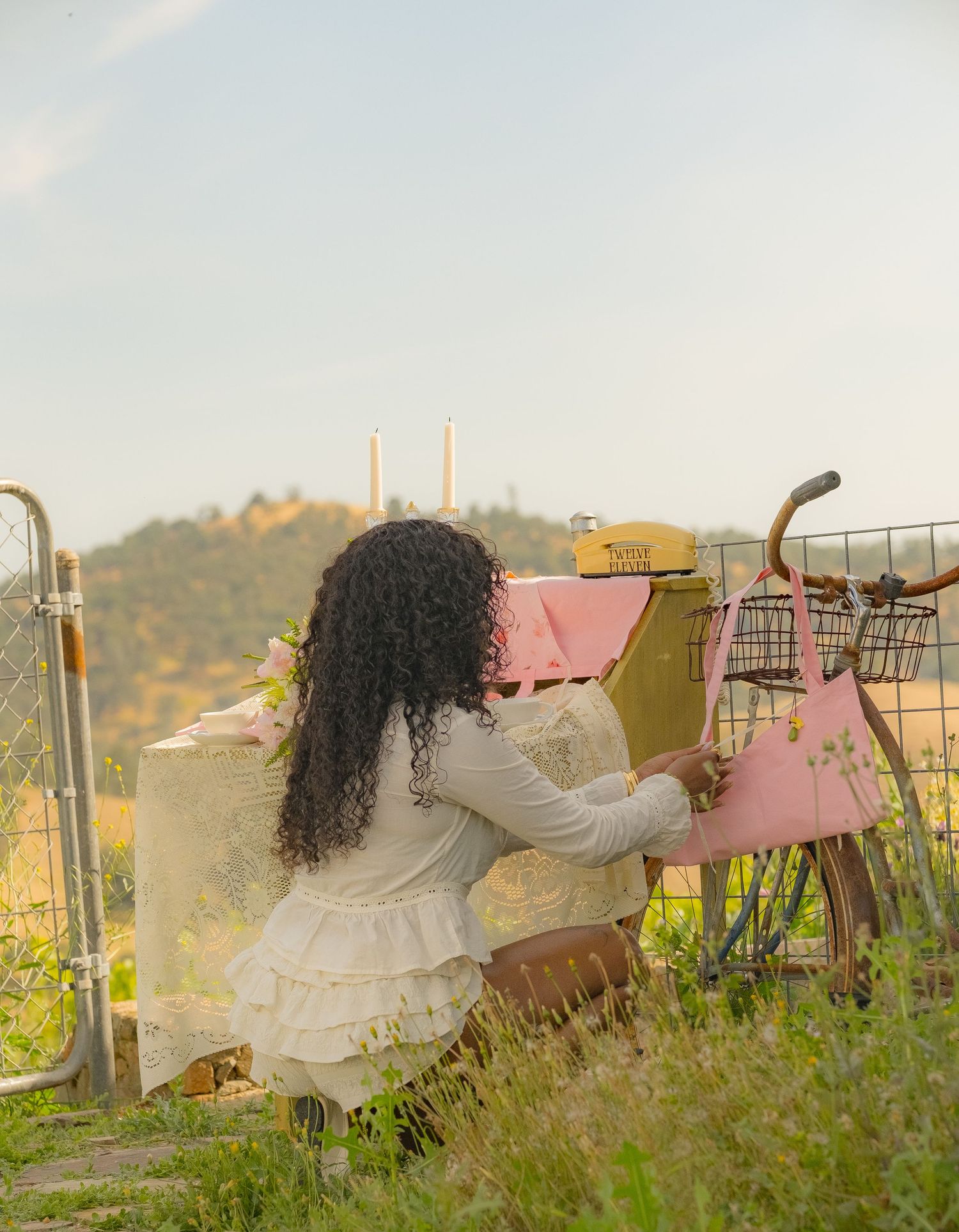 Young woman with curly hair kneeling by a vintage yellow bike with pink bags in a scenic countryside setting.