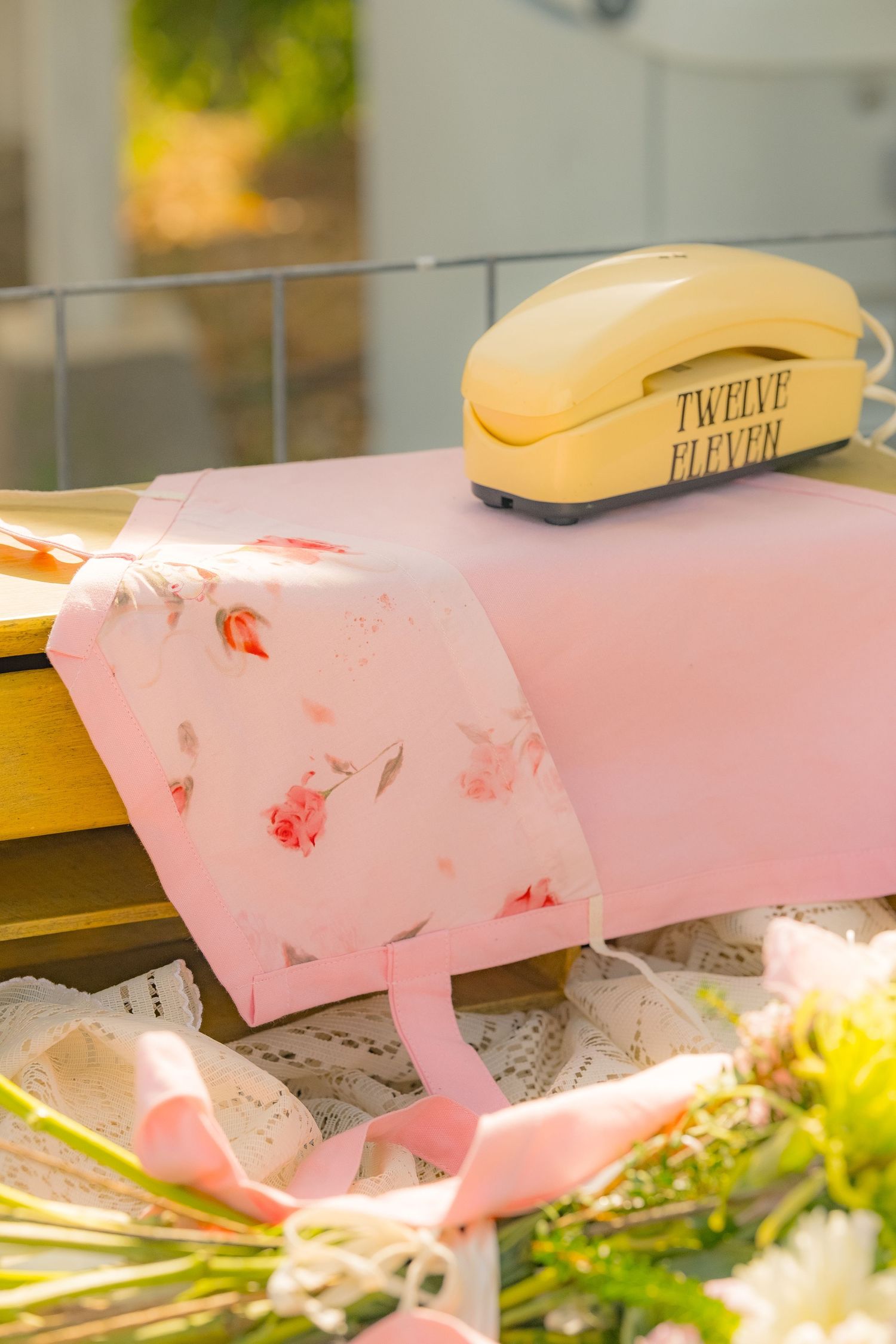 Pink floral tote bag on wooden bench with vintage yellow telephone labeled Twelve Eleven in sunny outdoor setting.