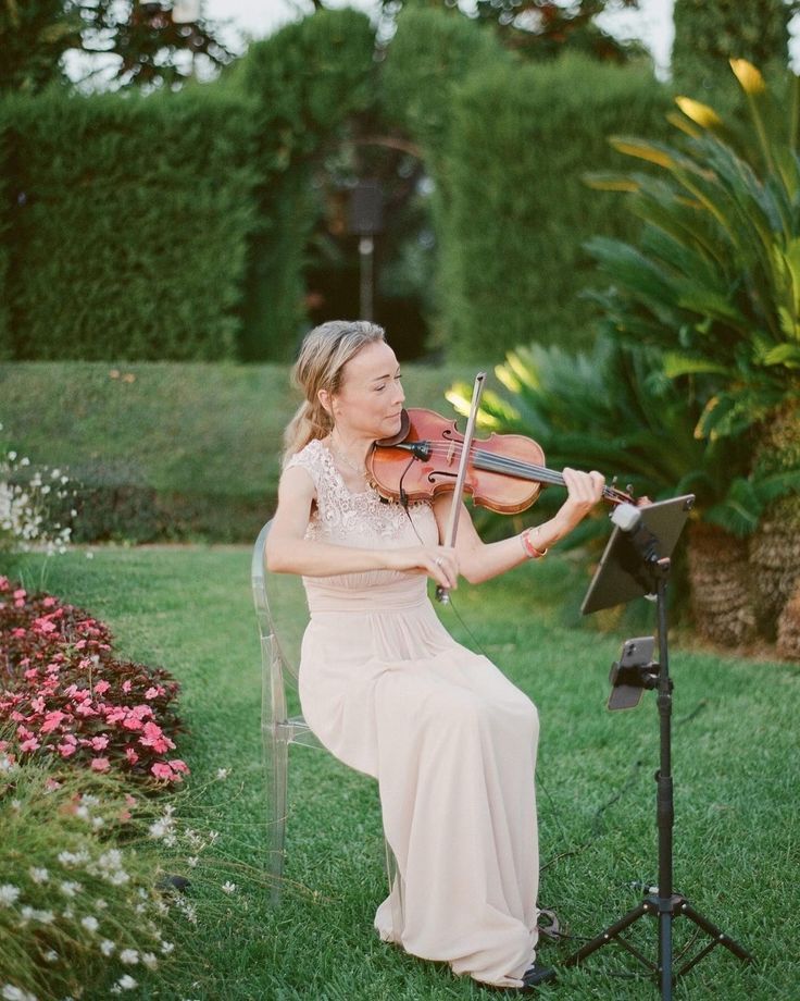 Elegant violinist in a blush gown performing outdoors in a lush garden setting at a wedding ceremony.