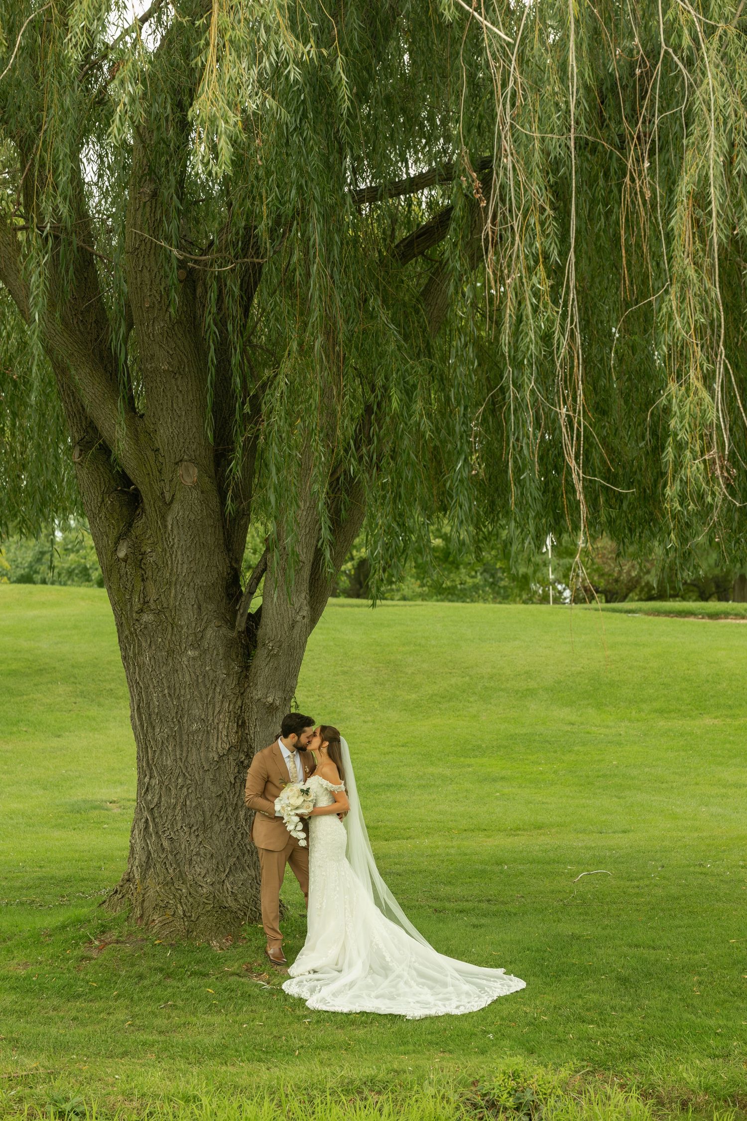 Bride in a white gown with a long veil and groom in a tan suit embrace beneath a large weeping willow tree on a green lawn.
