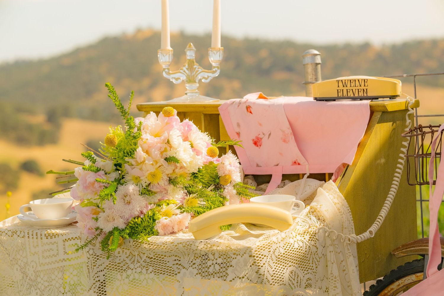 Romantic outdoor wedding tablescape with pink roses, lace cloth, candles, and rolling hills backdrop.