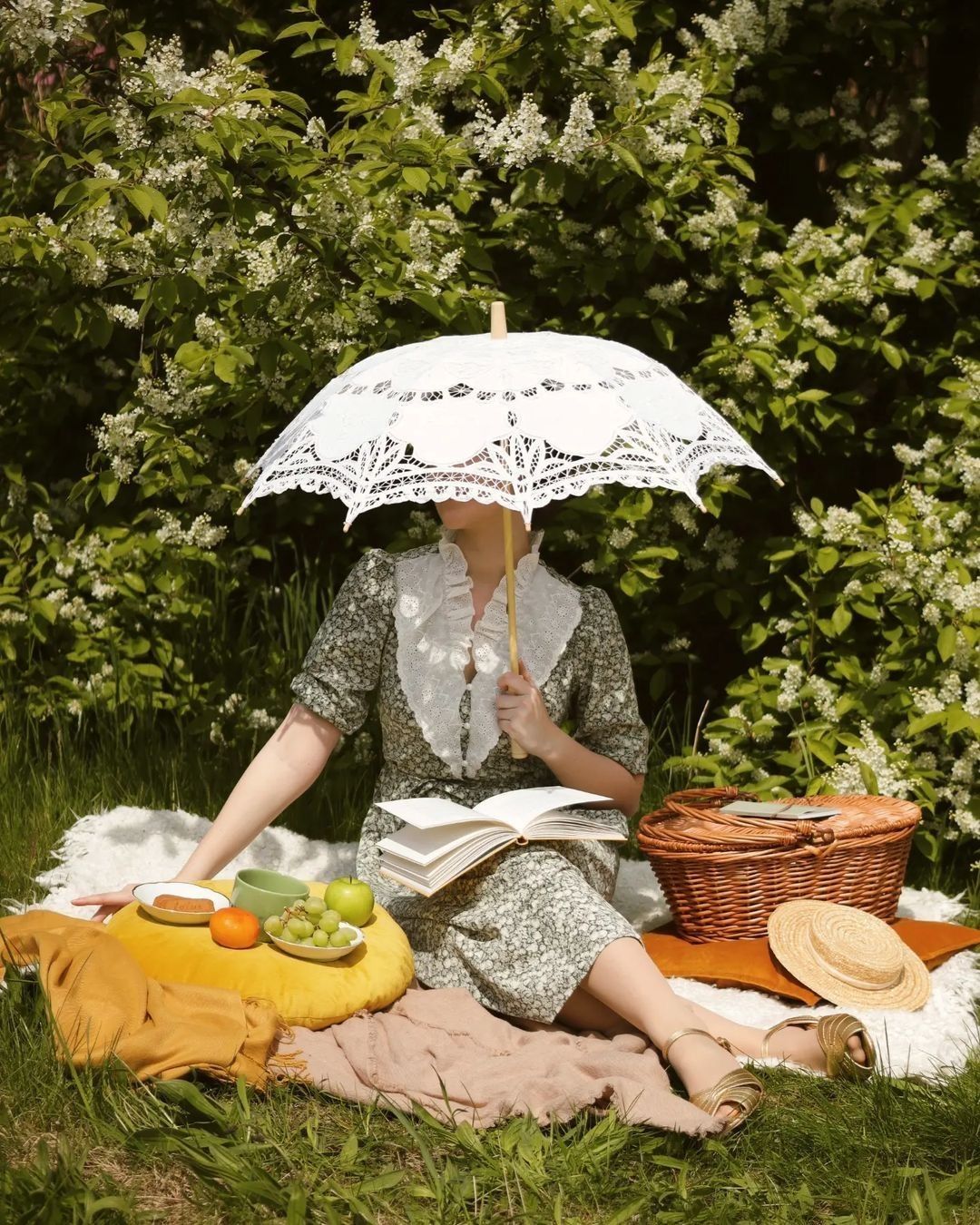 Woman reading a book at a garden picnic under a white lace parasol, surrounded by fruit and a wicker basket.
