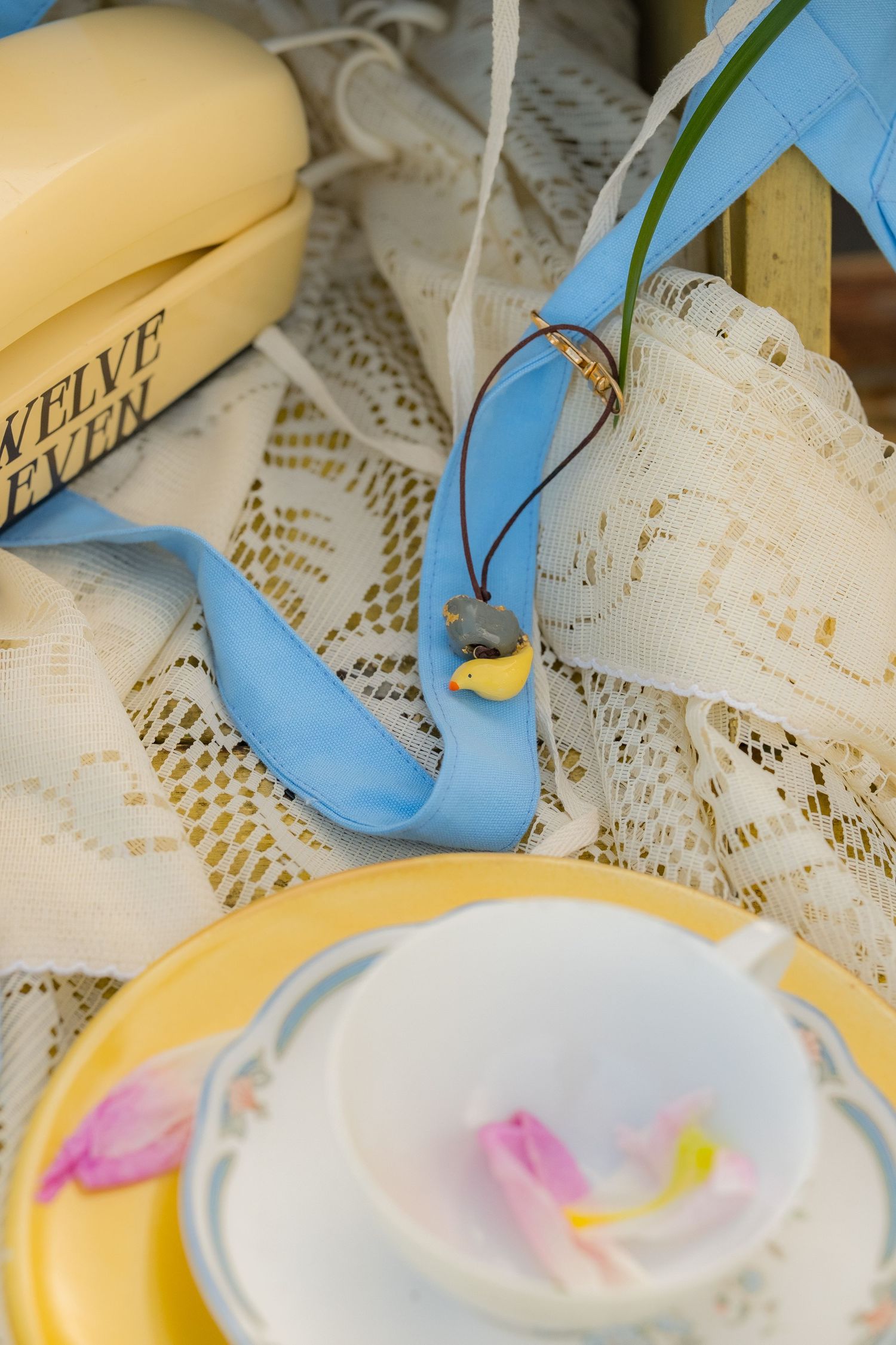 Vintage tea cup with flower petals on yellow saucer, lace fabric, blue ribbon, and retro yellow phone on books.