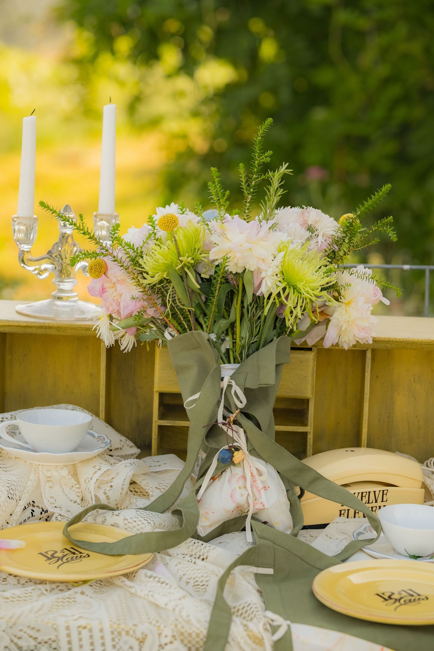 Elegant outdoor table setting with yellow plates, floral centerpiece, candles, and lace tablecloth in a garden.