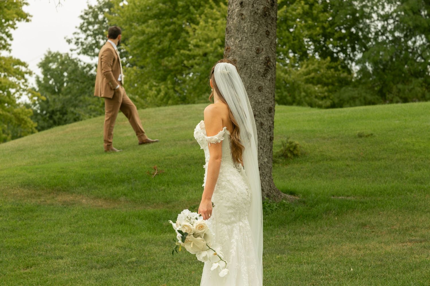 Bride in an off-shoulder lace wedding gown and long veil holds a white bouquet while the groom in a tan suit walks up a grassy hill.
