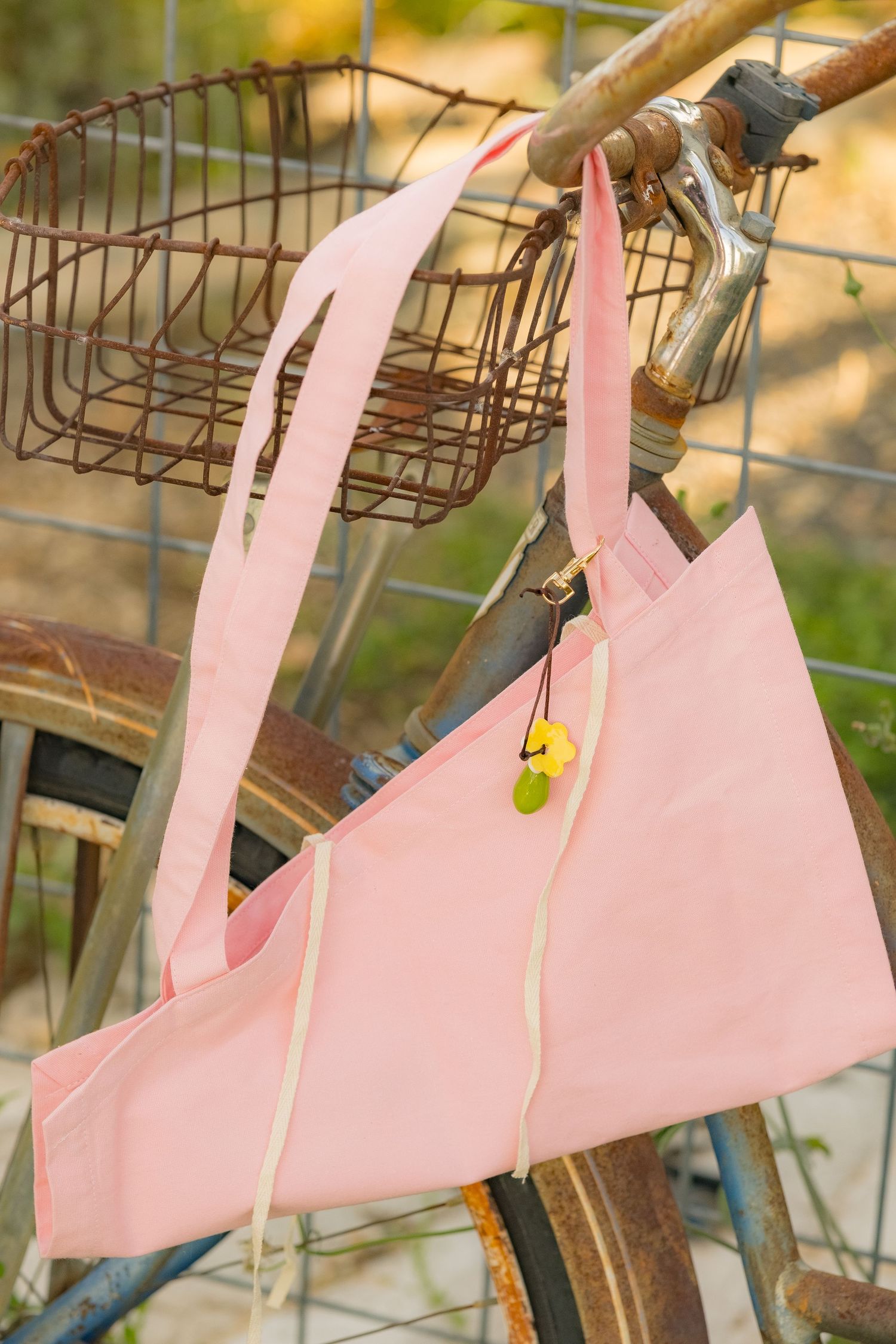 Pink tote bag with yellow charm hanging on rusty vintage bicycle with wire basket outdoors.