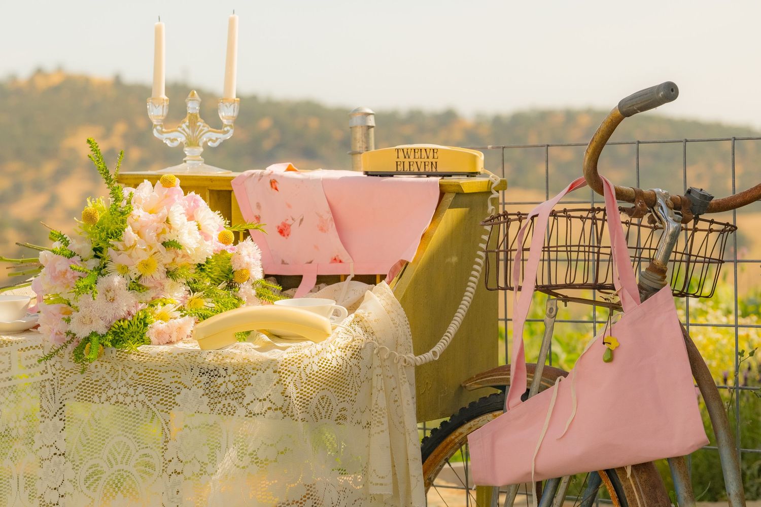 Romantic outdoor table setting with lace tablecloth, pink accents, candles, floral bouquet, and vintage bicycle in soft light.