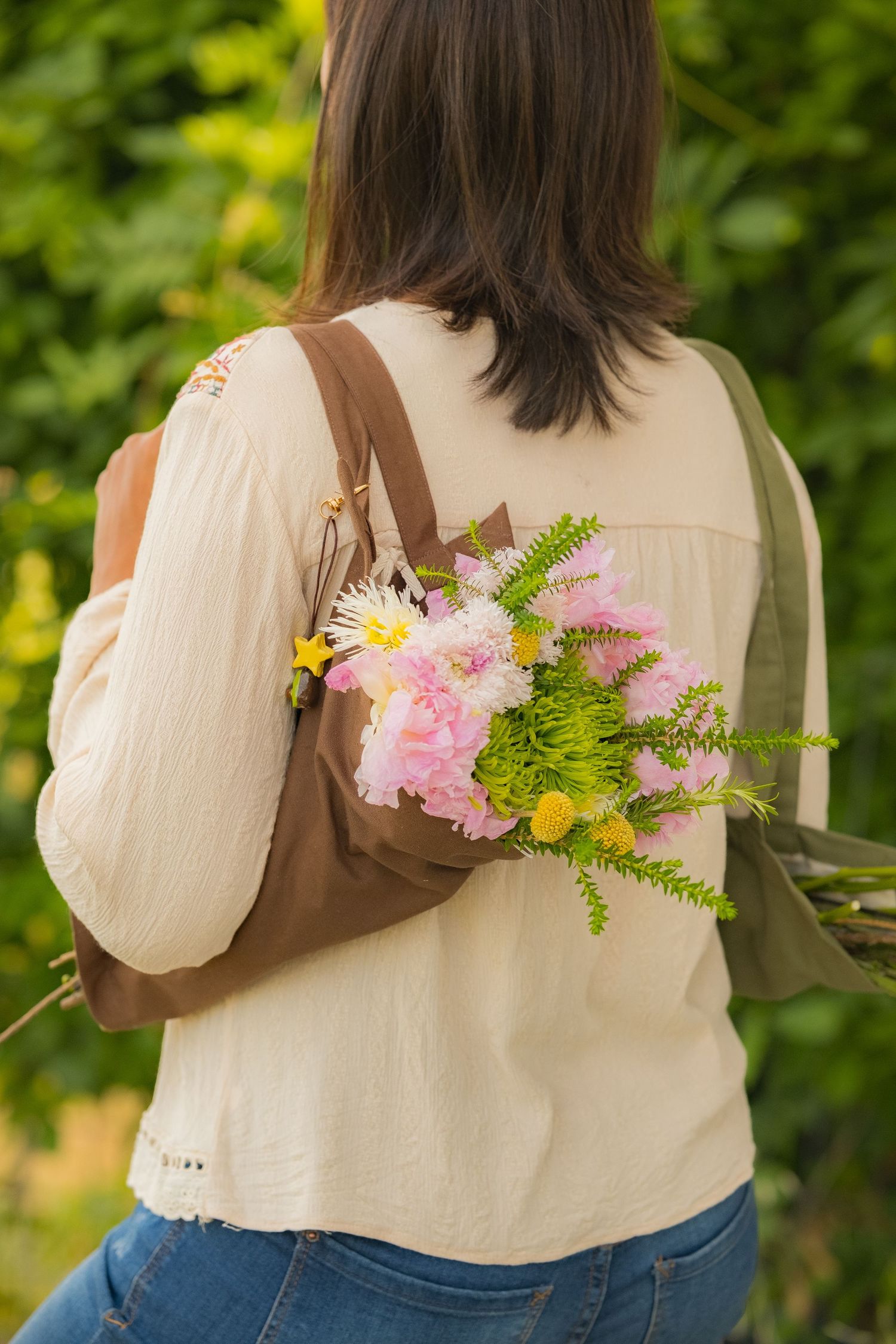 Woman seen from behind holding a bouquet of pink and yellow wildflowers in a brown backpack outdoors.
