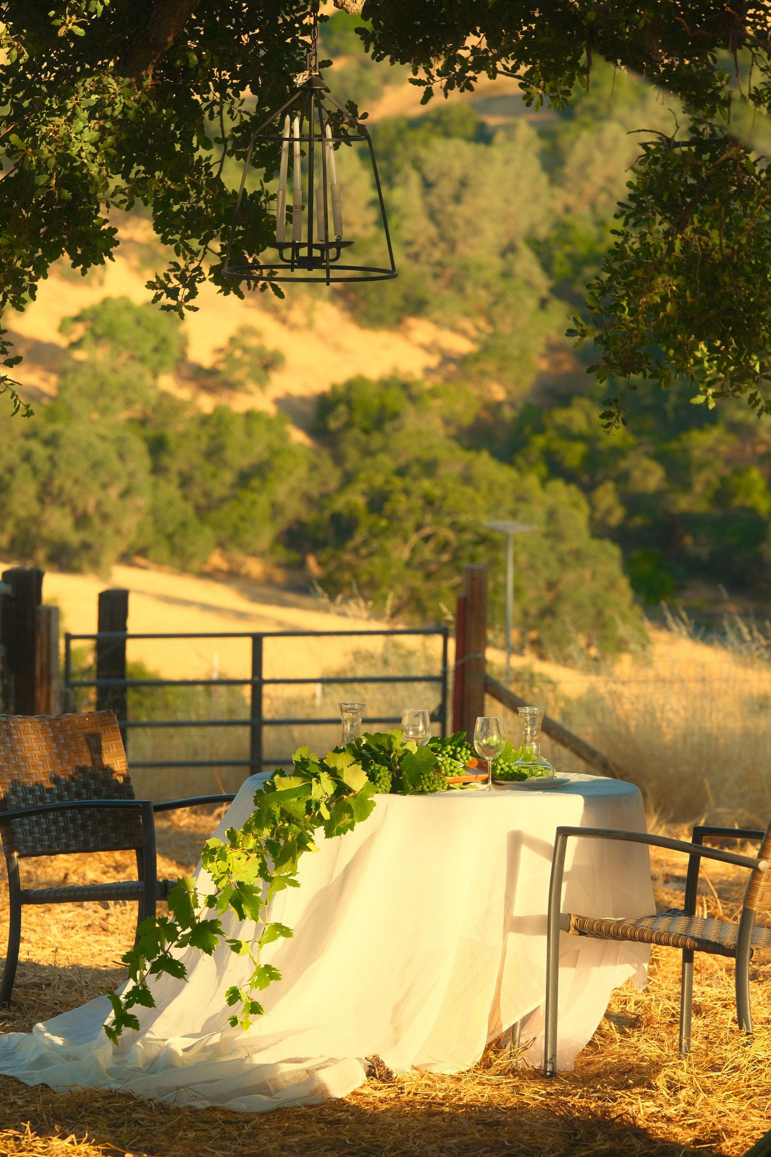 Outdoor wedding setup with white tablecloth and chairs against a scenic countryside backdrop at sunset.