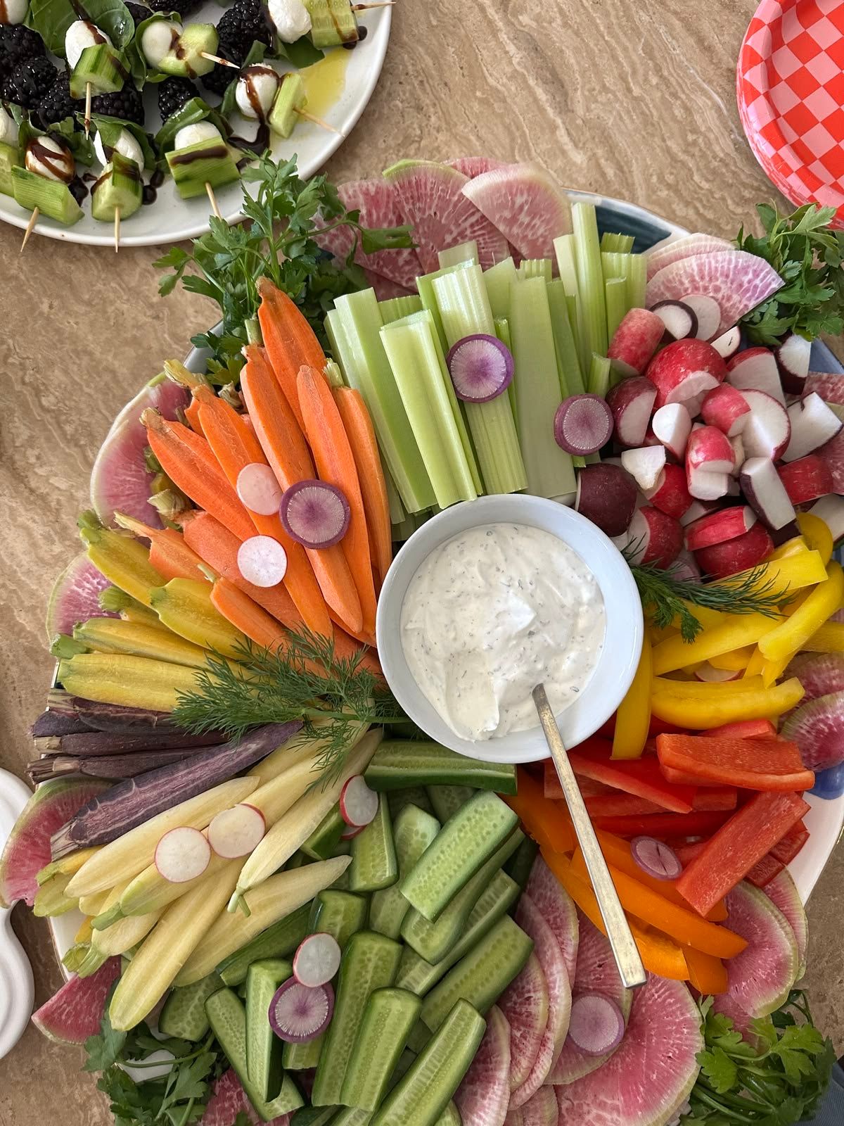Colorful vegetable crudité platter with celery, carrots, radishes, peppers, and creamy white dip in center bowl.