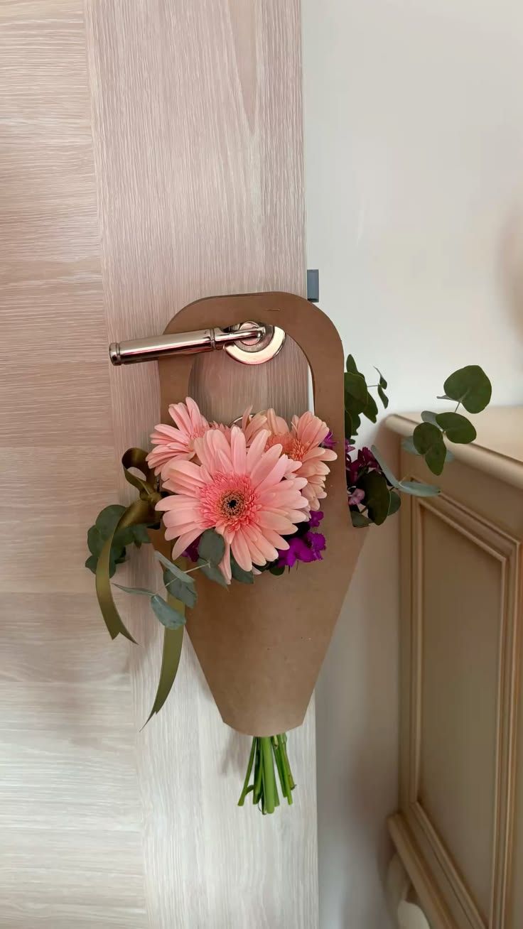 Pink gerbera daisy bouquet in kraft paper cone hanging on a door handle with purple flowers and eucalyptus greenery.