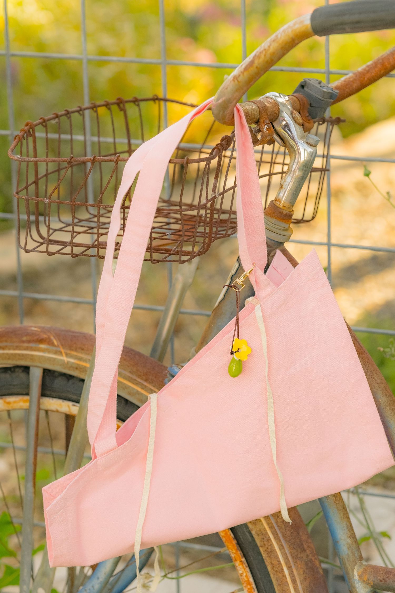 Pink canvas tote bag with yellow charms hanging from rusty vintage bicycle handlebar outdoors.