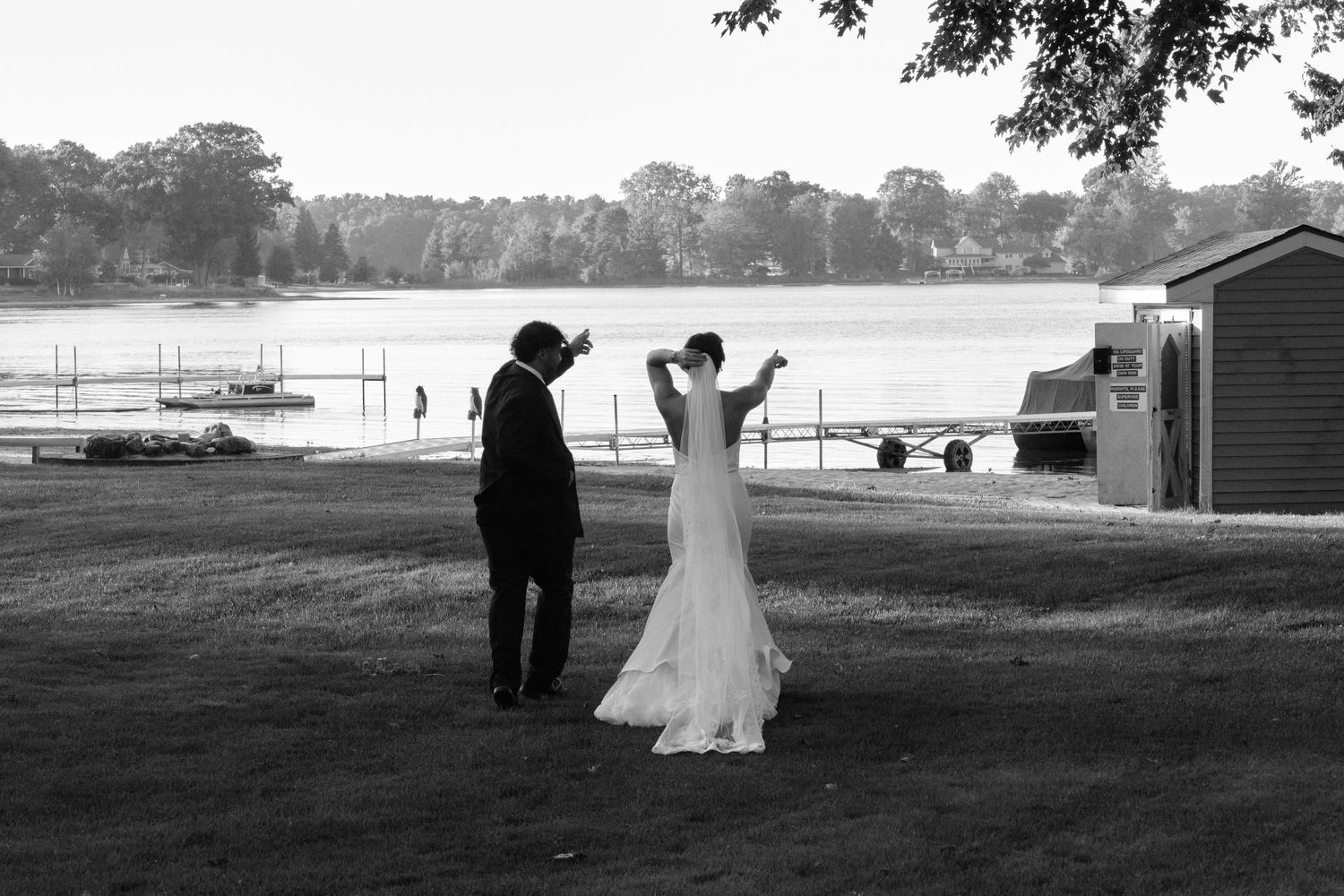 Black and white photo of two figures in formal attire standing on a lawn overlooking a lake with boats in the distance.