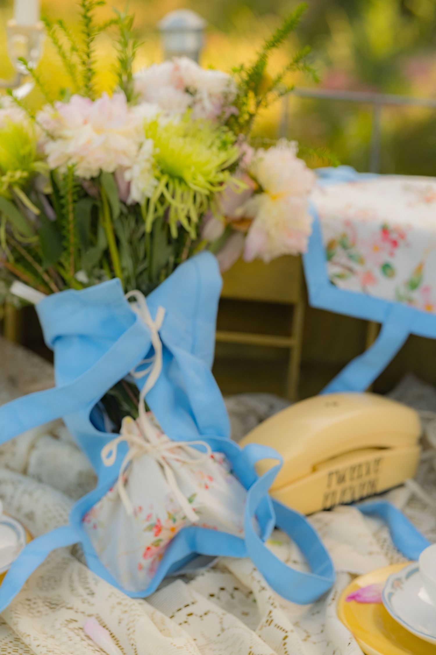 Vintage table setting with blue ribbon, floral bouquet, yellow rotary phone, and lace tablecloth outdoors.