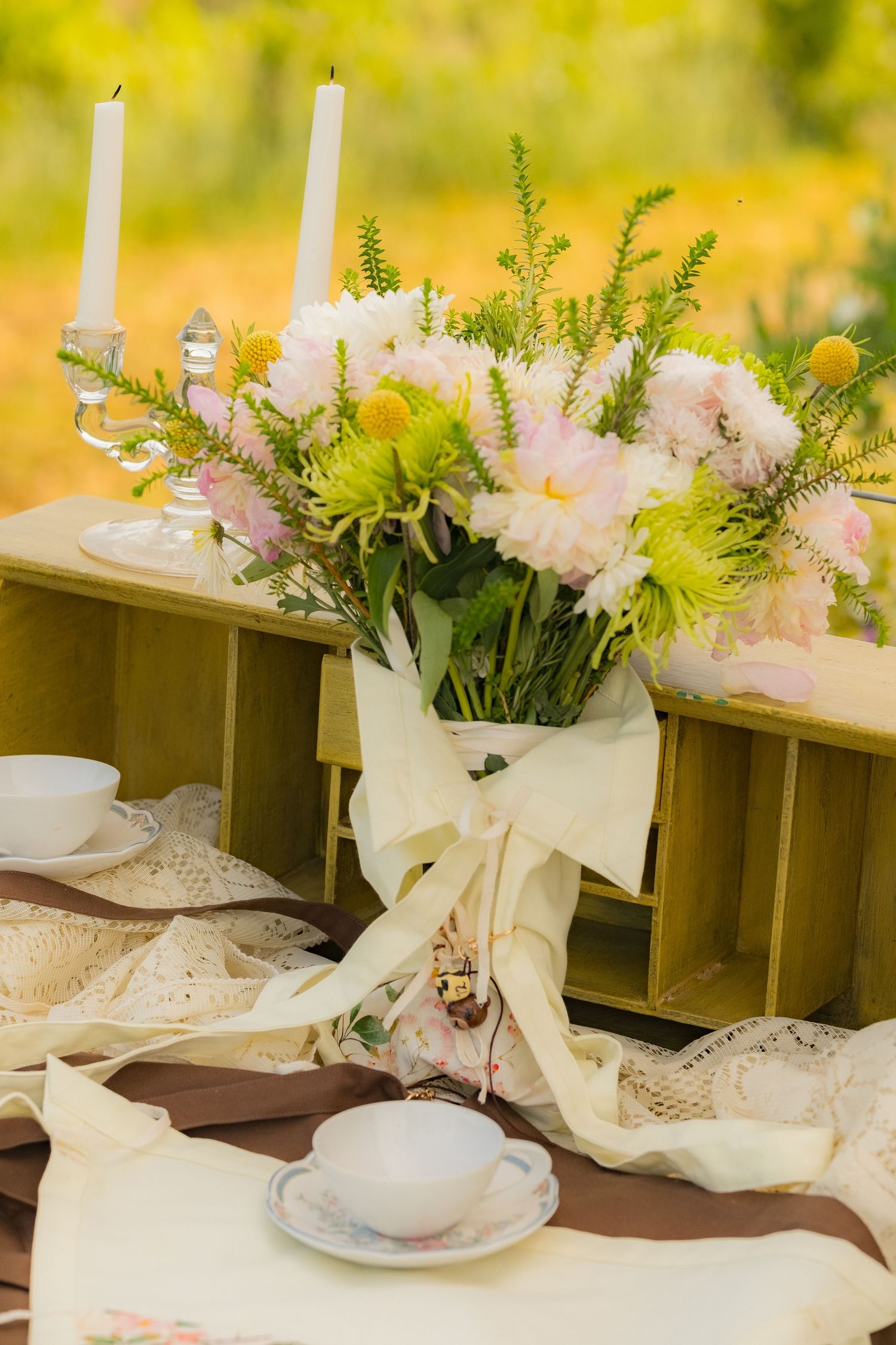 Elegant outdoor table setting with pink and yellow flowers in a bouquet, white candles, and fine china teacups on lace cloth.