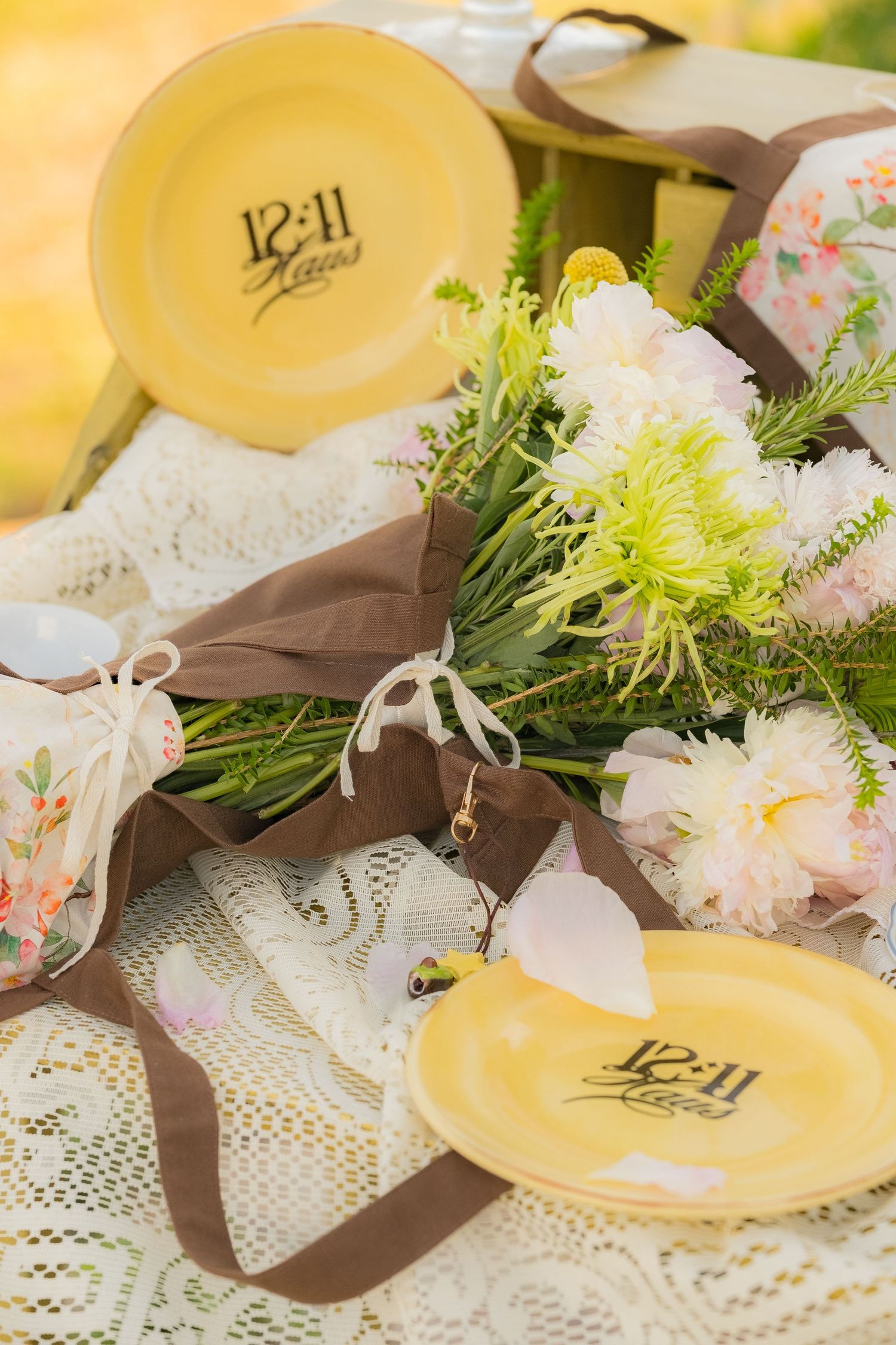 Yellow plates with 12:11 Haus branding displayed on lace tablecloth with pink peonies and brown ribbon bouquet.