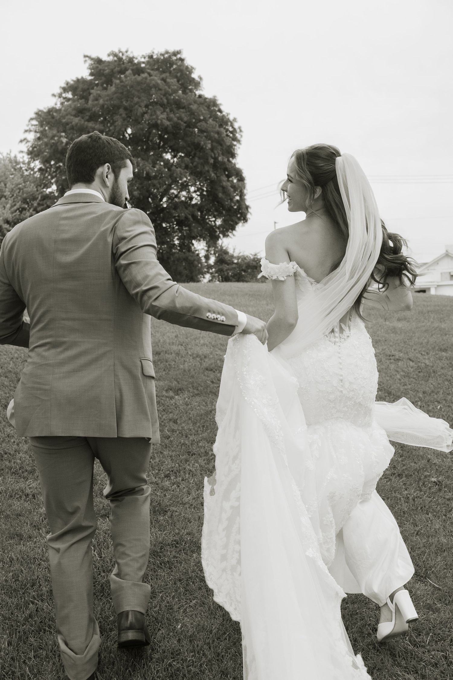 Black and white photo of a bride in a lace gown and veil holding her train as she walks hand in hand with a groom across a grassy lawn.