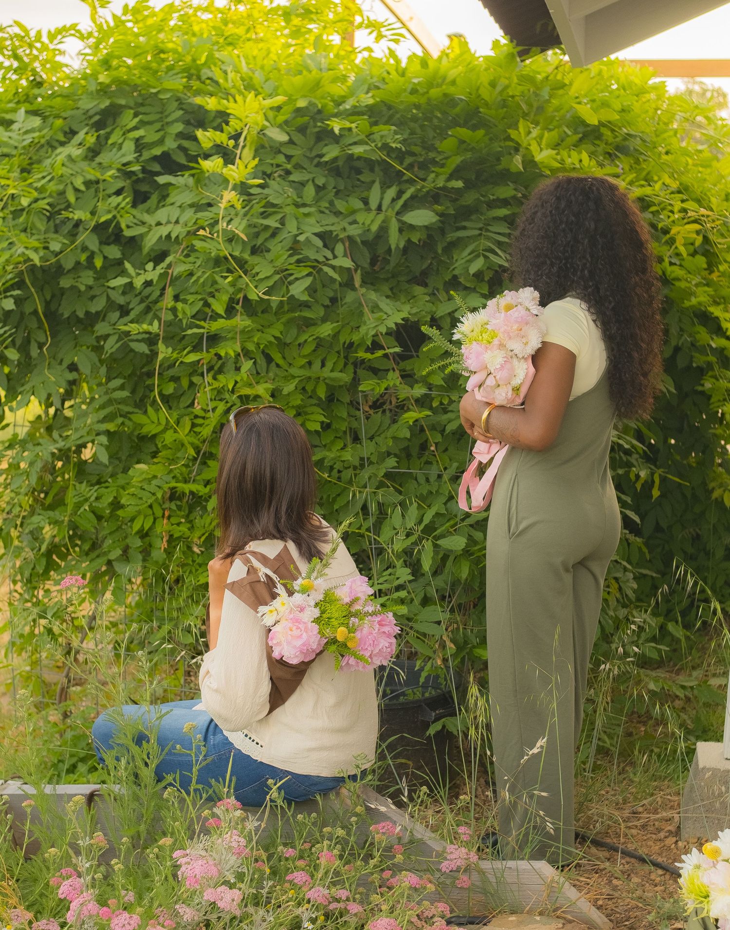 Two women holding pink flower bouquets stand near a lush green hedge in a garden setting.