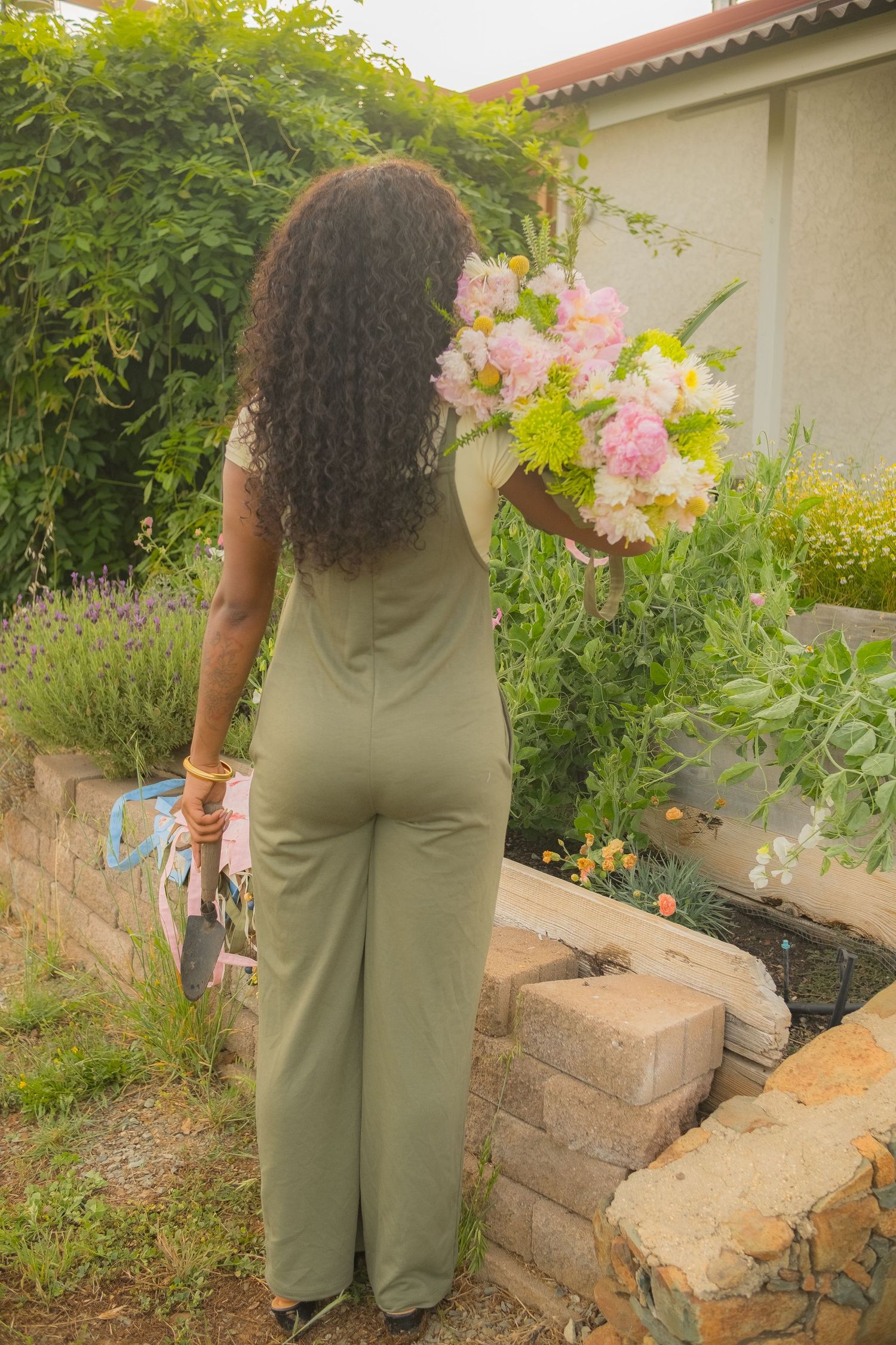 Woman in olive green jumpsuit holding a bouquet of pink and white flowers in a lush garden setting.