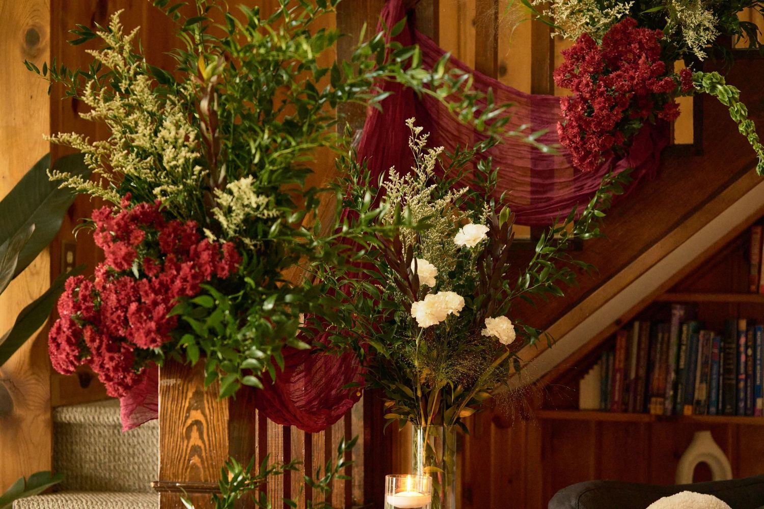 Elegant floral arrangement with deep red blooms and greenery displayed on wooden staircase with bookshelves.