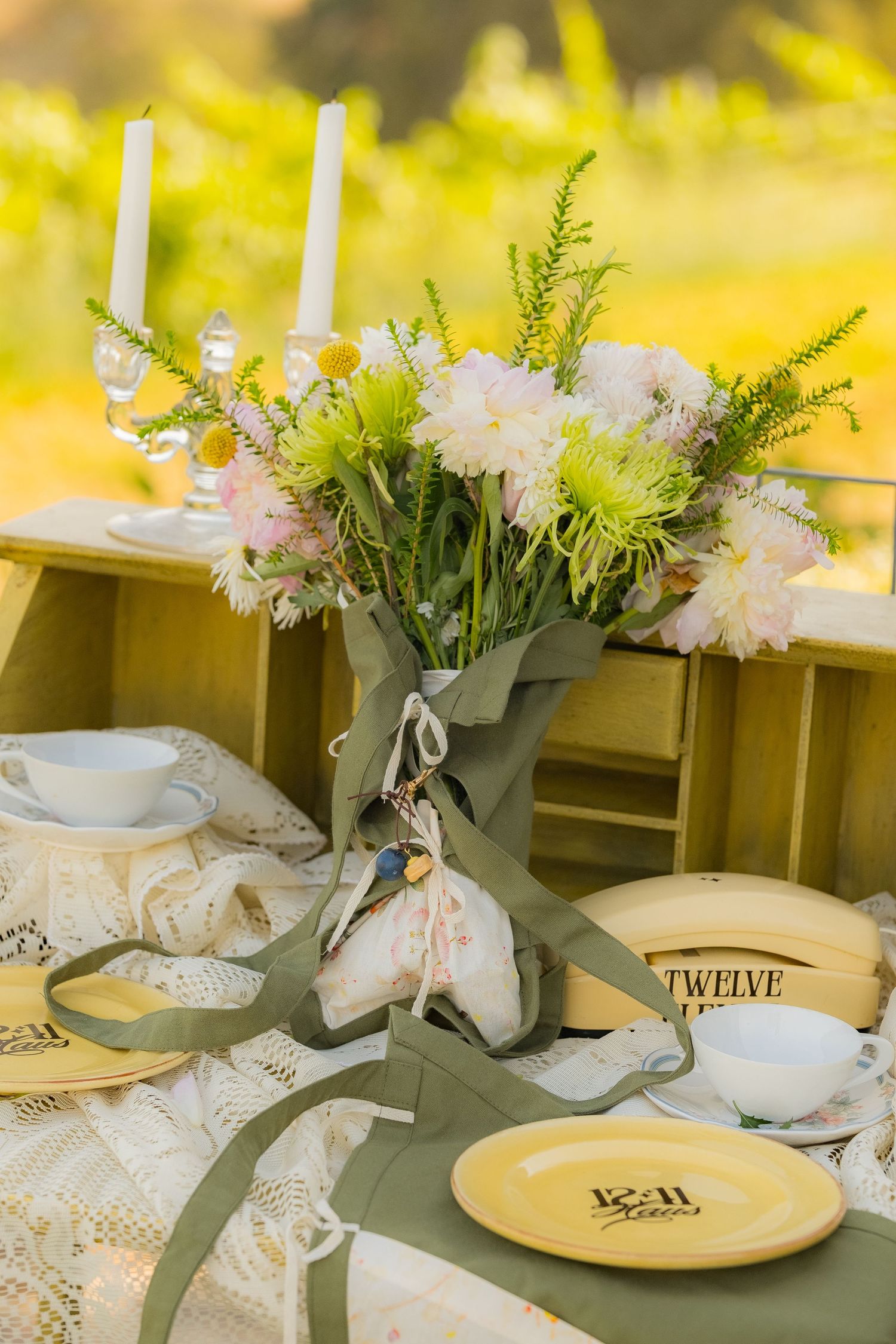 Elegant outdoor table setting with floral centerpiece, yellow plates, candles, and lace runner in a garden.