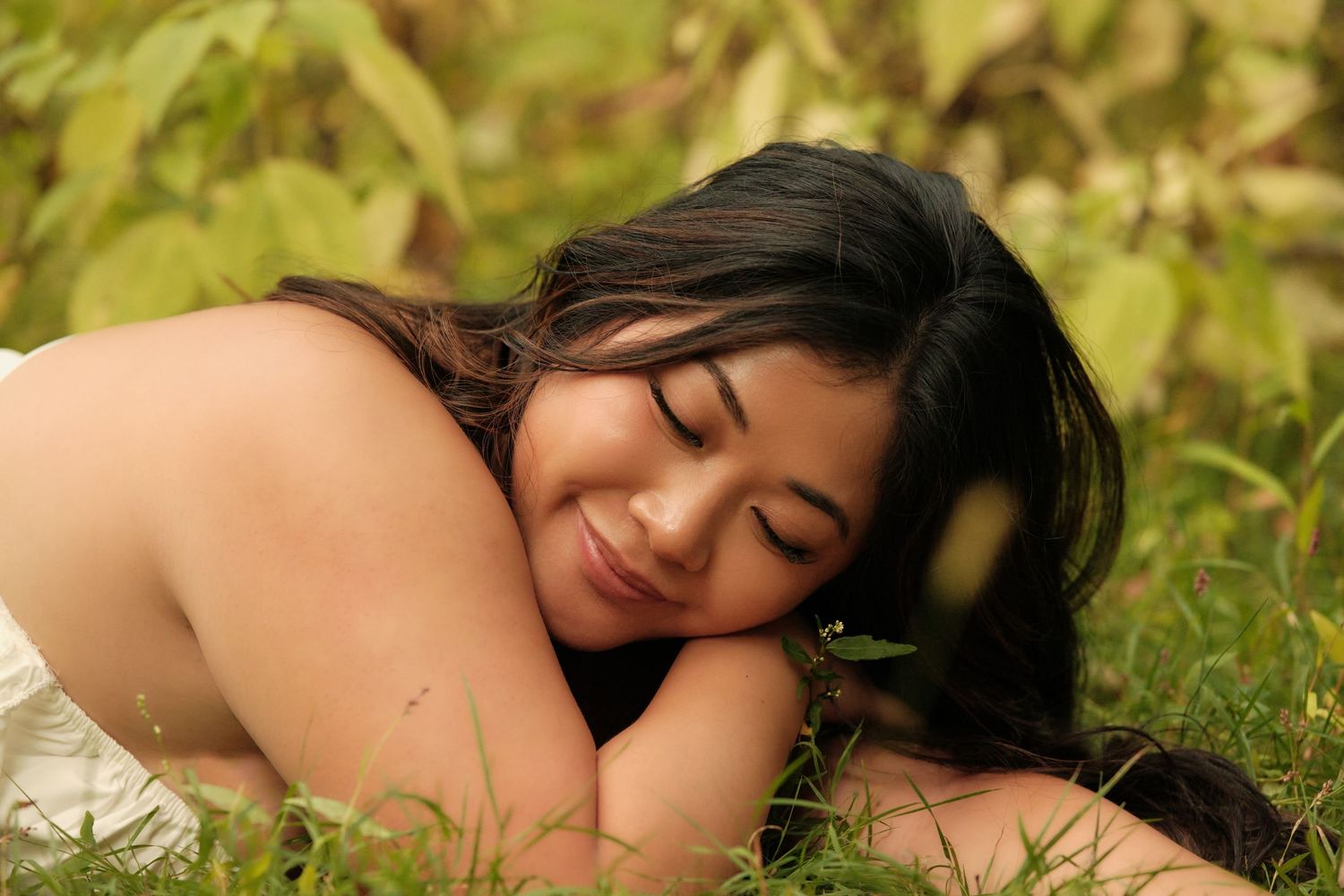 A person with long dark hair lies peacefully in green grass surrounded by autumn leaves.