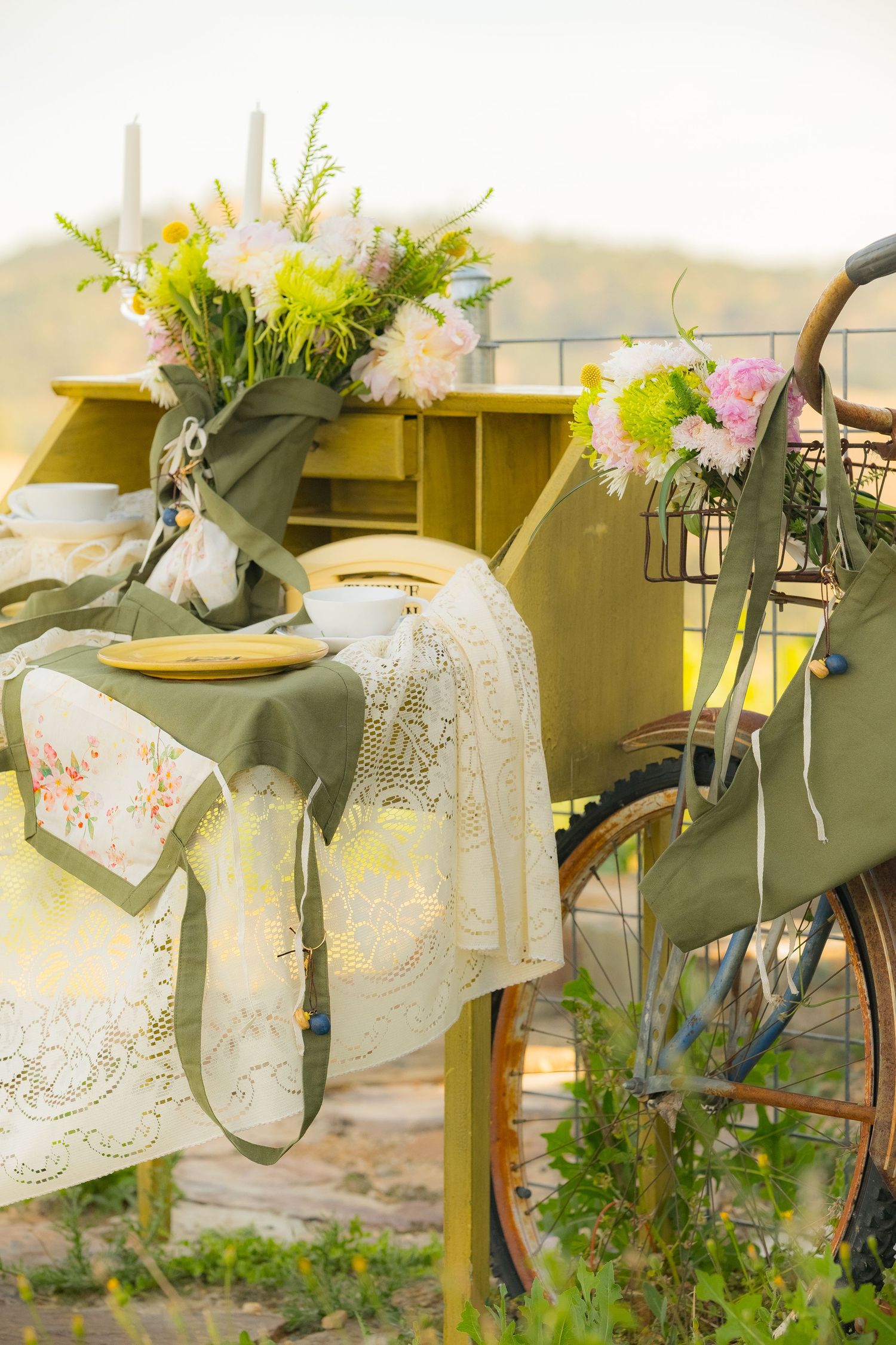 Vintage outdoor table setting with lace cloth, yellow plates, flowers, and a rustic bicycle adorned with blooms.