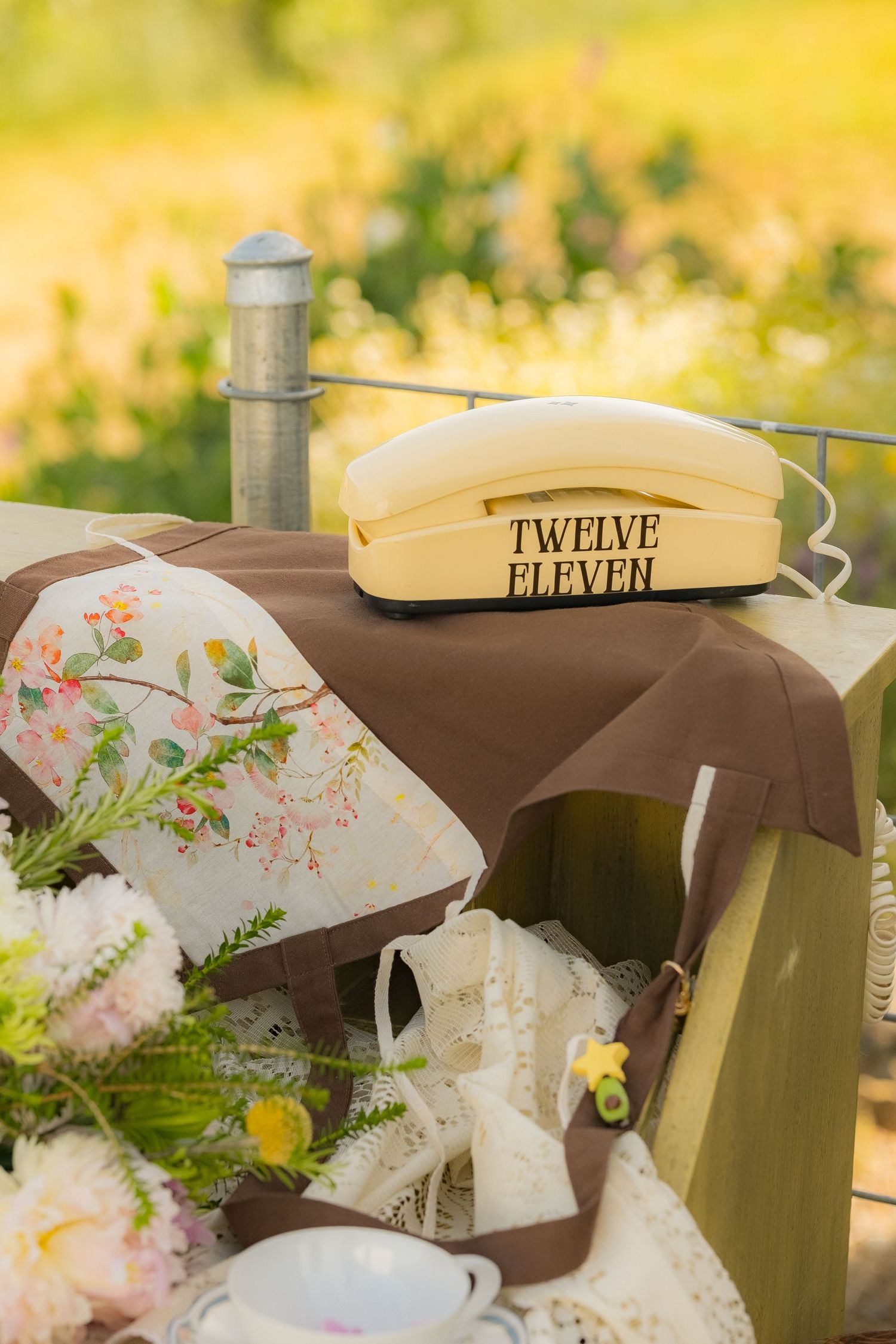 Vintage cream telephone labeled Twelve Eleven on brown cloth with floral pillows and flowers outdoors.