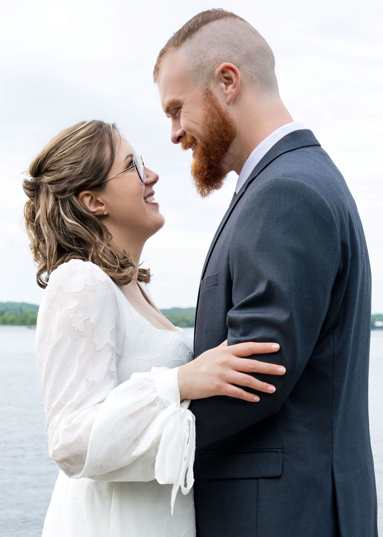A bride in white dress and groom in gray suit share a romantic moment by the water on their wedding day.