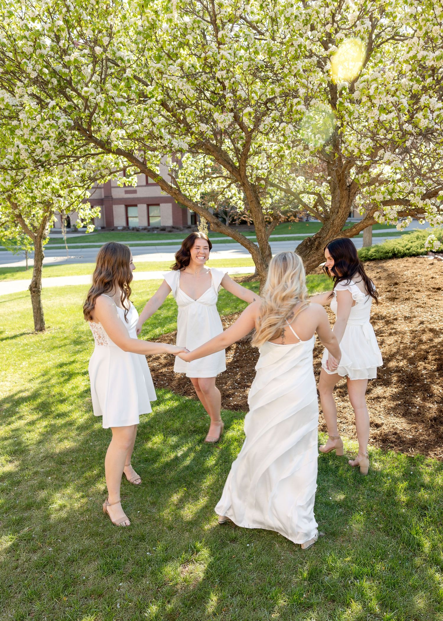 Bride in white gown dancing with bridesmaids in white dresses under blooming trees on green lawn.