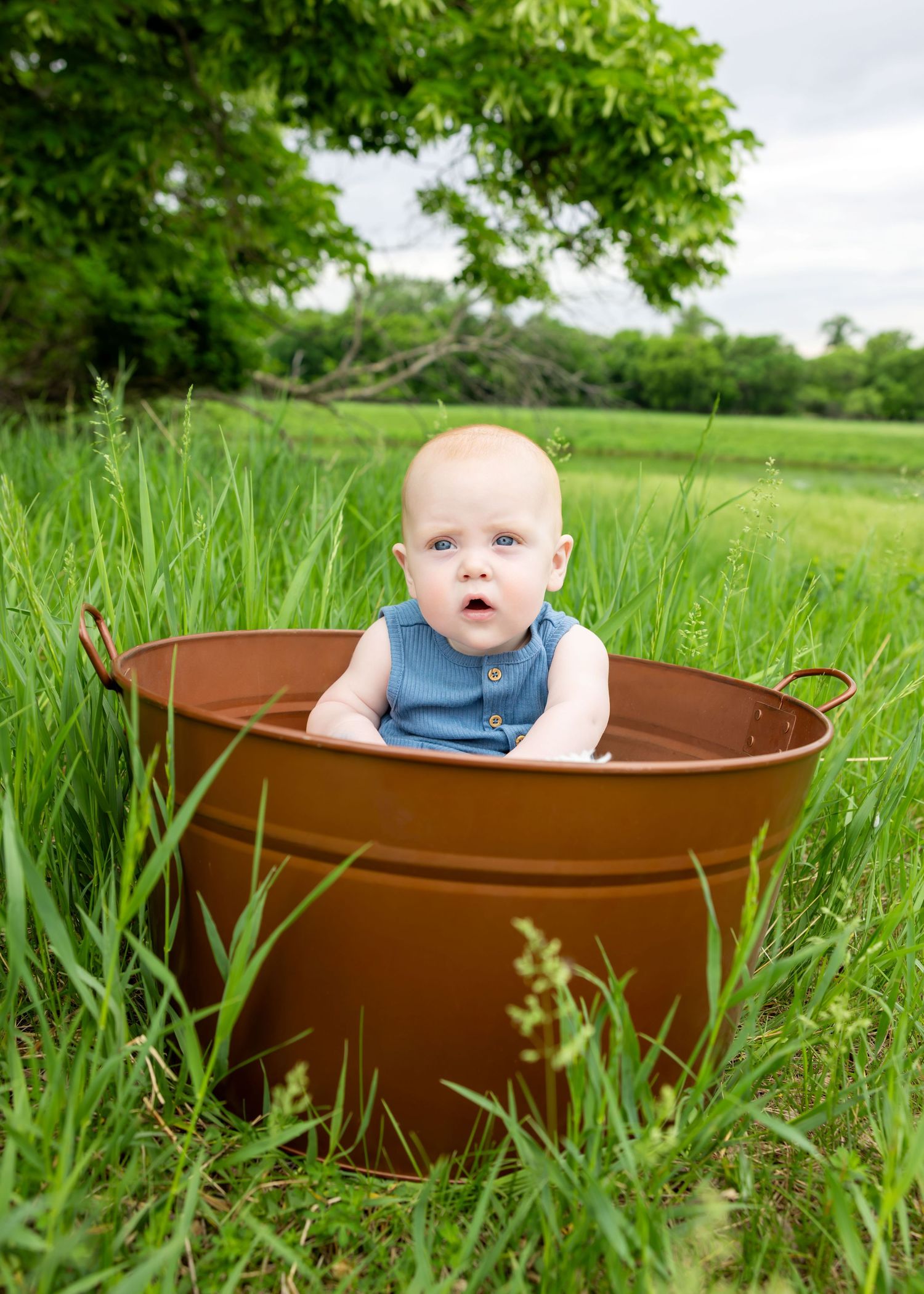A cute baby in a blue romper sits inside a rustic brown metal tub placed in a lush green grassy field.