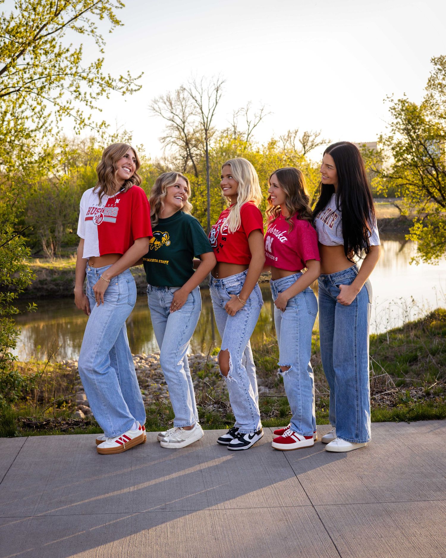 Five young women in colorful crop tops and jeans pose together outdoors near a pond with lush green trees.