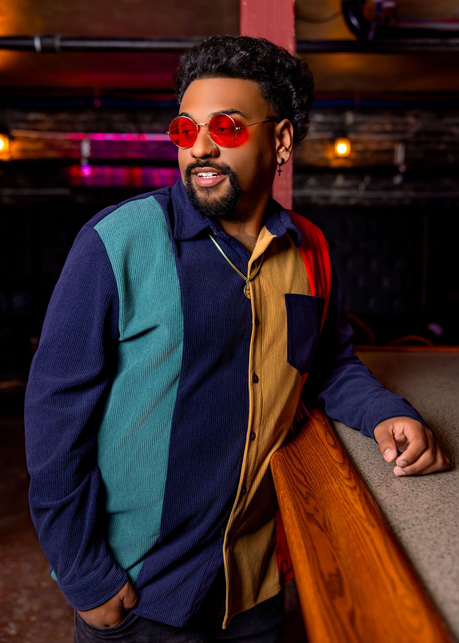 Man in colorblock jacket and red round sunglasses posing confidently in a stylish indoor setting.