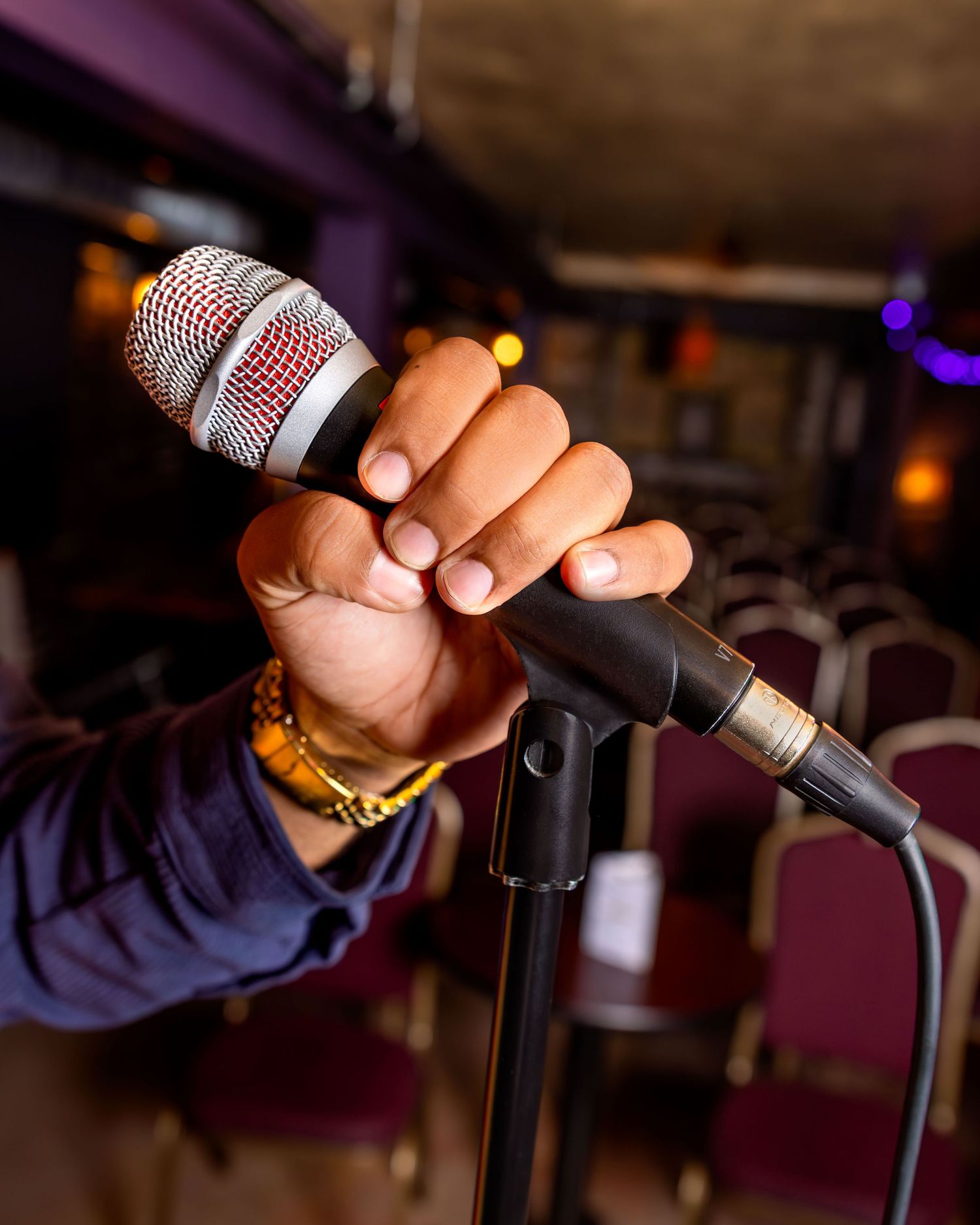 A hand gripping a microphone on a stand in a dimly lit venue with empty red chairs in the background.