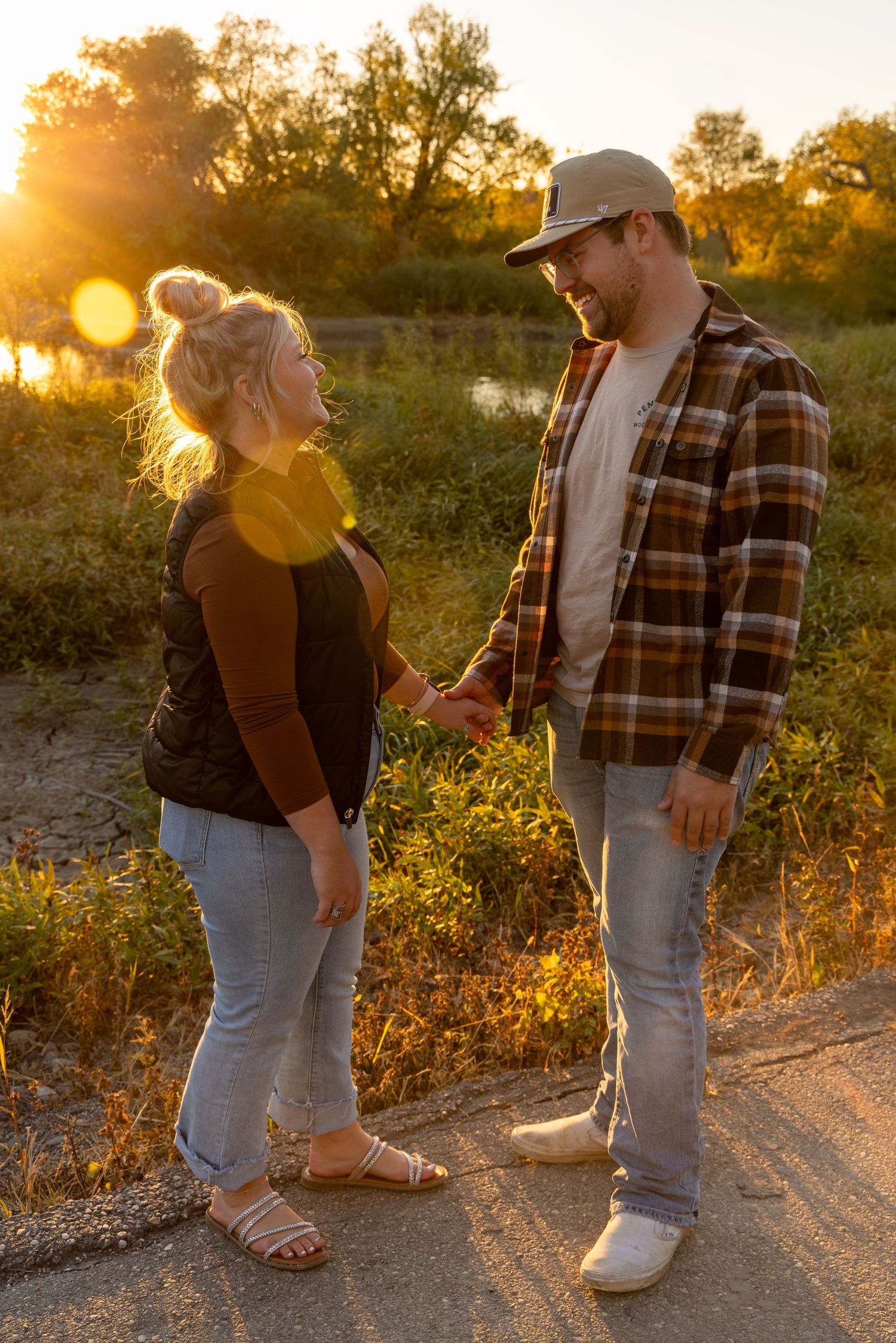 A couple stands together on a dirt path during golden hour wearing casual outdoor attire.