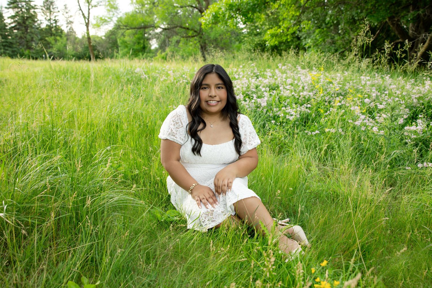 Young woman in white lace dress sitting in lush green meadow with wildflowers on a sunny day.