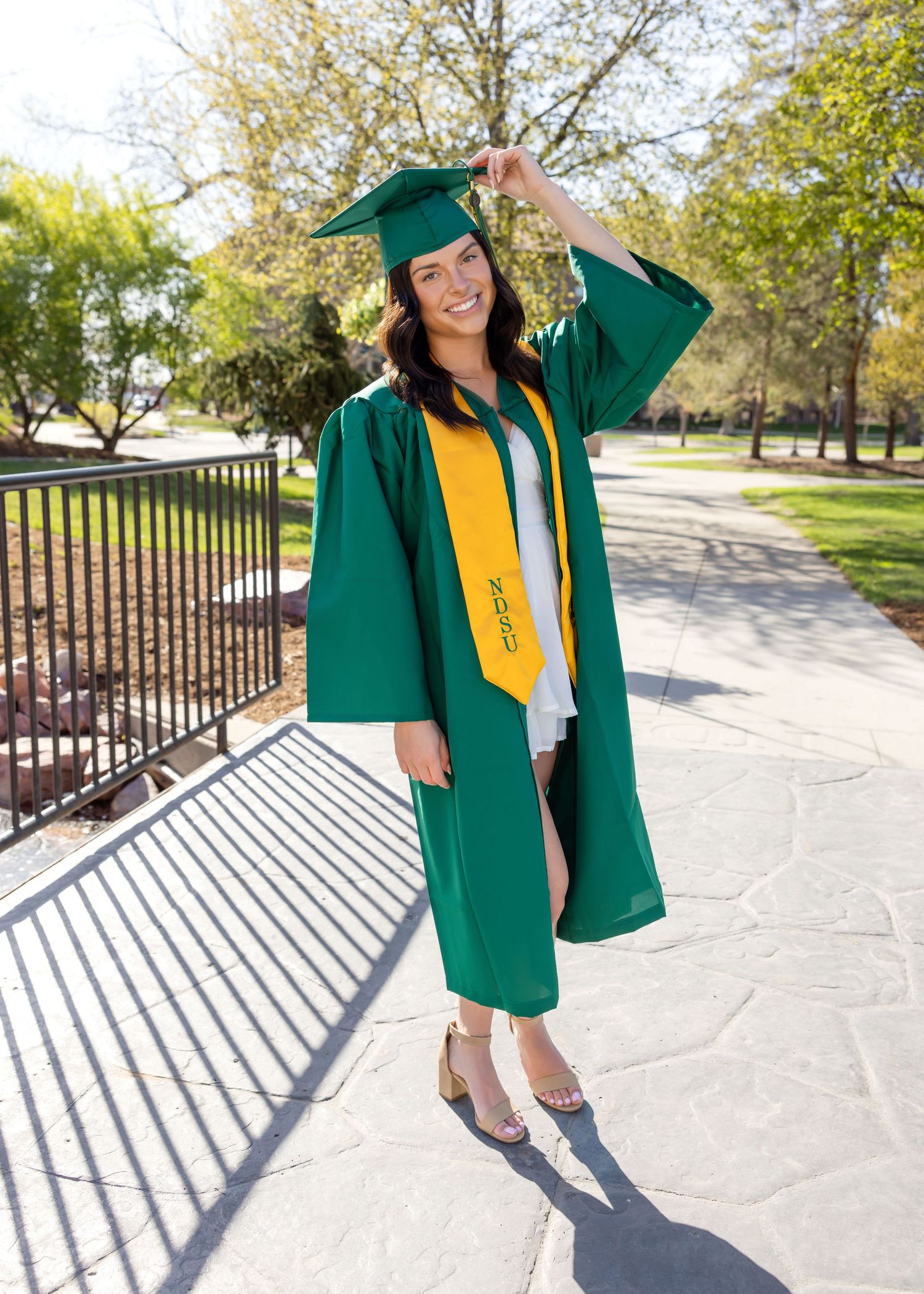 Smiling graduate in green cap and gown with gold stole poses on sunny campus walkway.