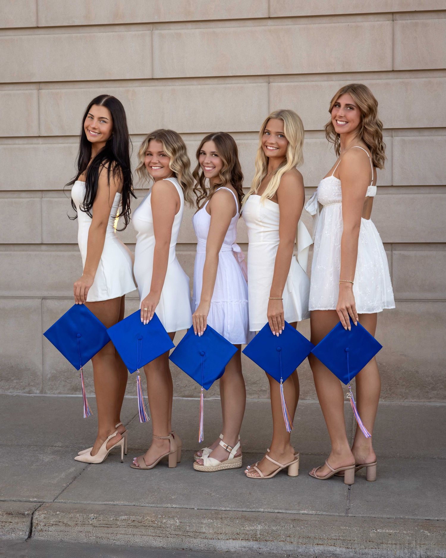 Five women in white dresses holding blue graduation caps pose together outdoors celebrating their graduation.