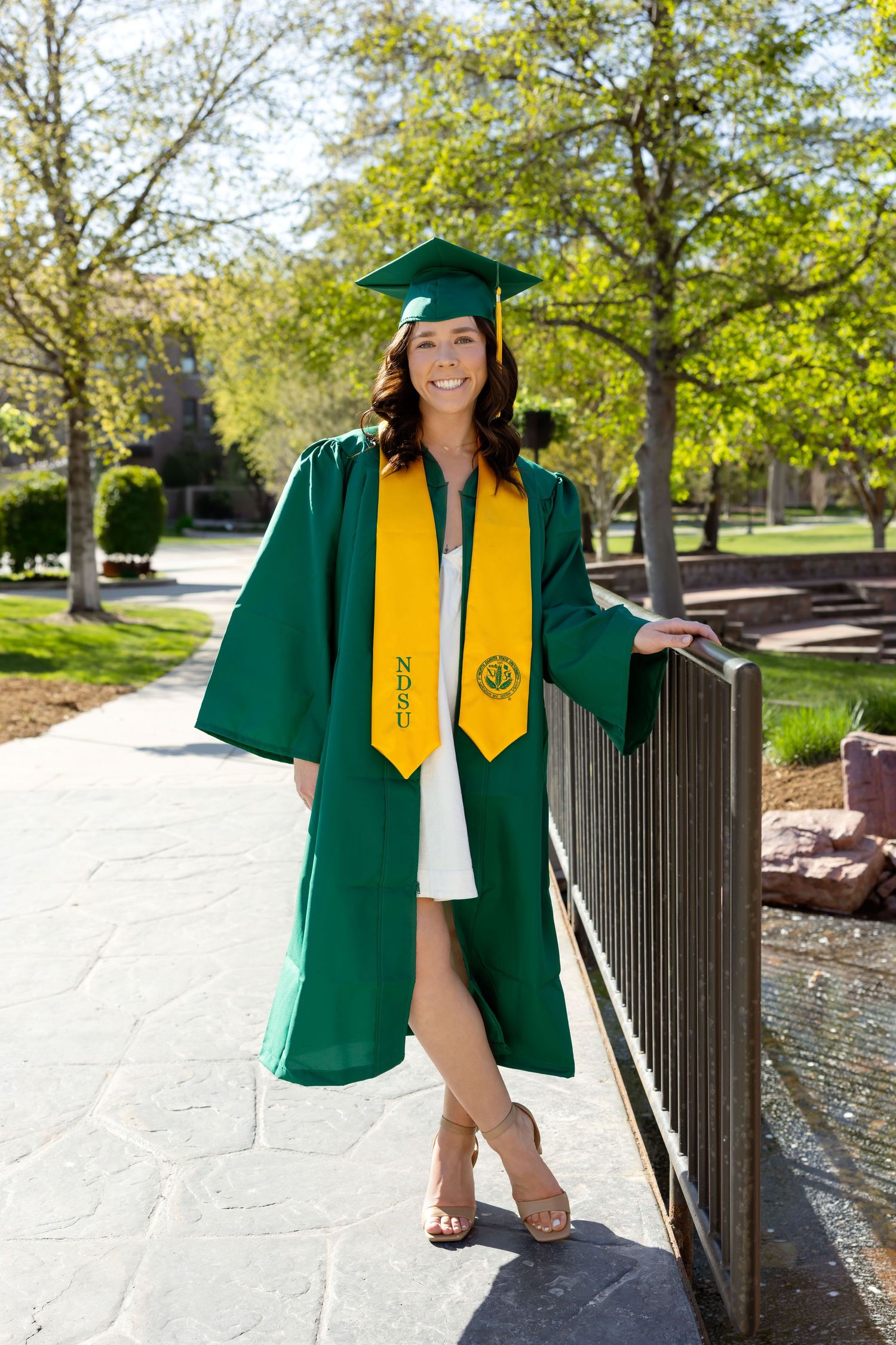 Smiling female graduate in green cap and gown with gold NDSU stole poses on a sunny campus walkway.