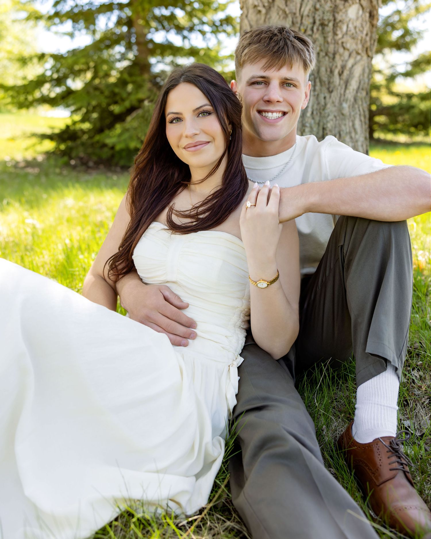 A smiling couple in wedding attire sits together on green grass beneath a tree outdoors.