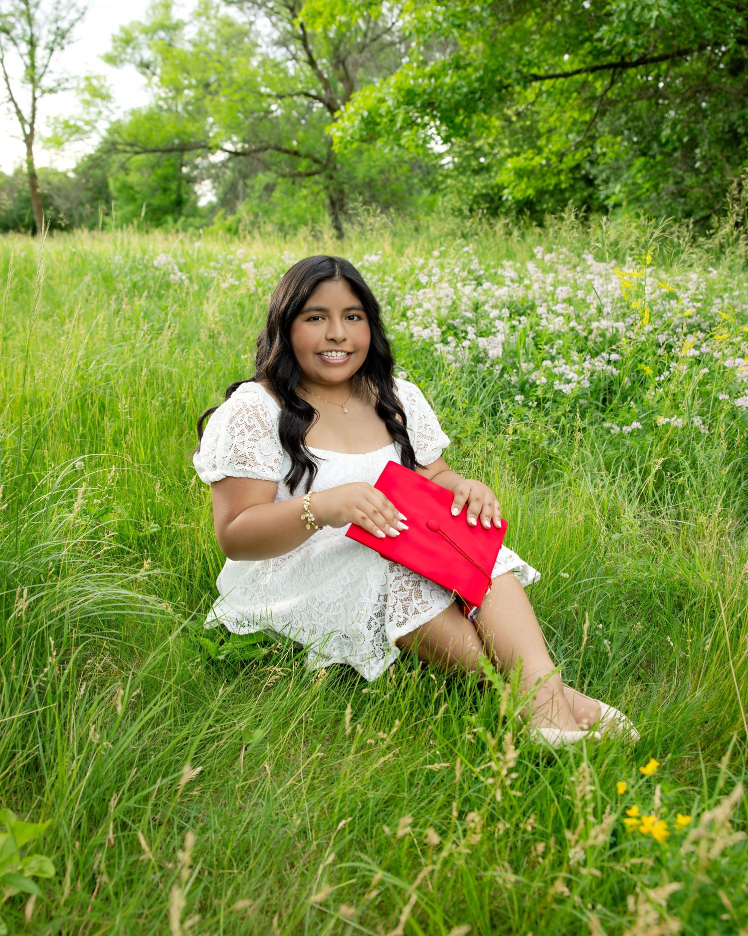 Young woman in white lace dress sits in green meadow holding red folder, smiling outdoors.